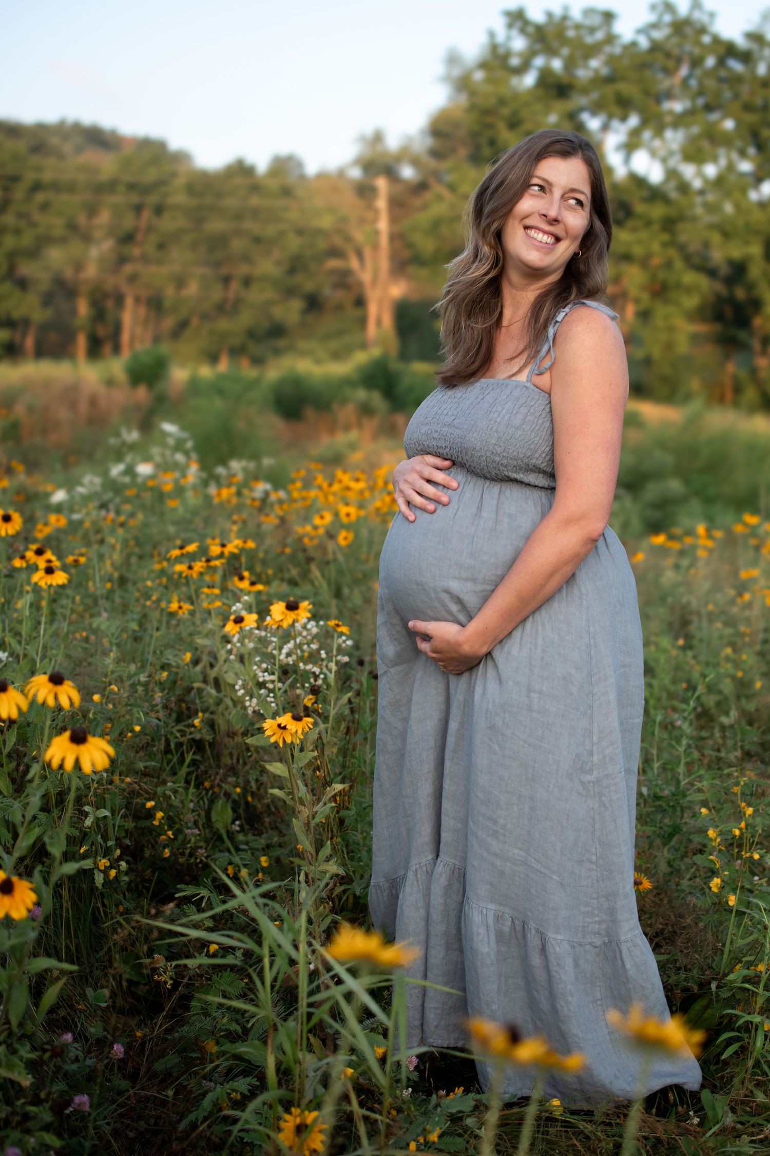 A pregnant woman in a gray dress stands in a wildflower field with yellow Black-eyed Susans blooming at sunset.