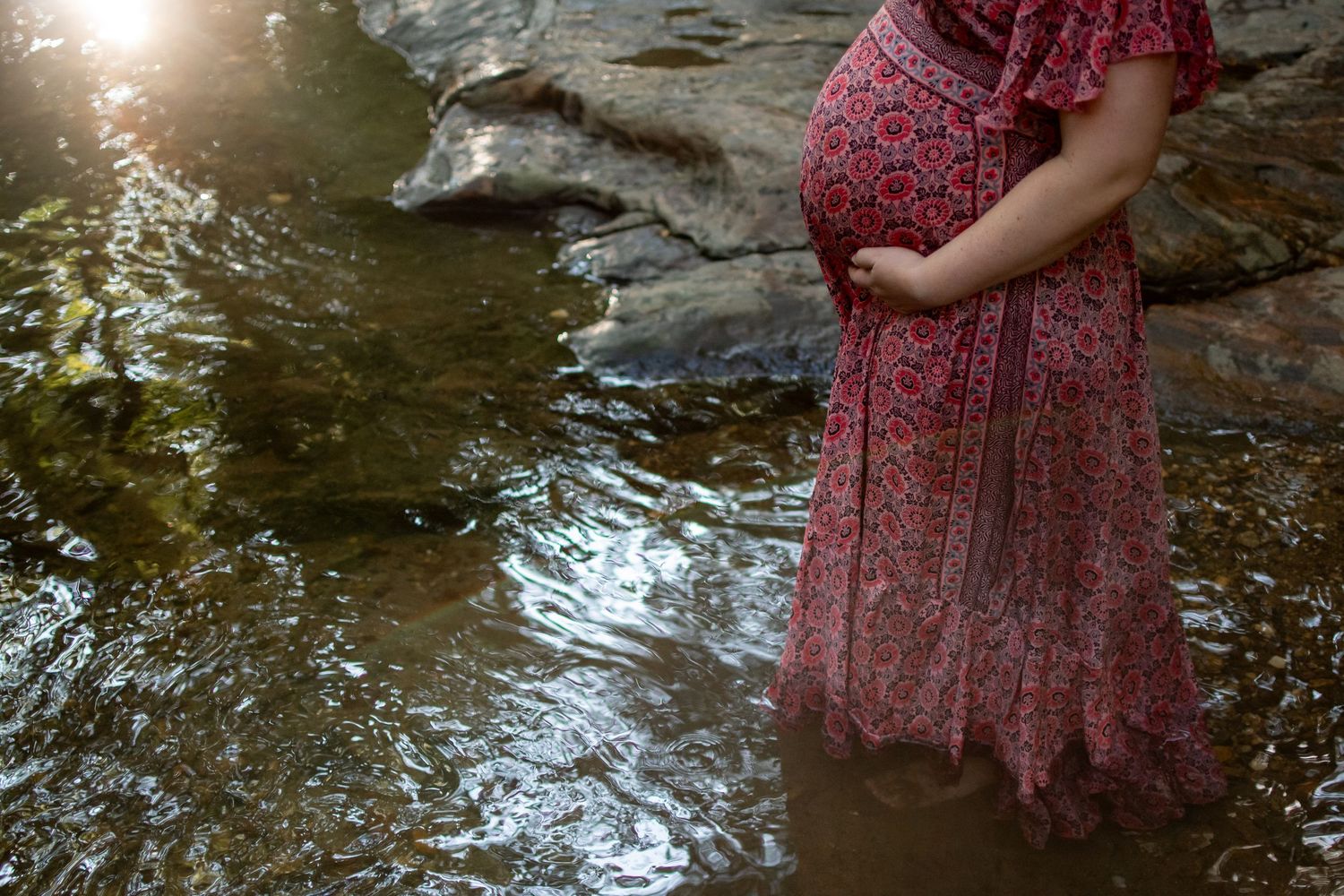 A person in a floral red dress stands in a shallow creek during a sunny summer day.