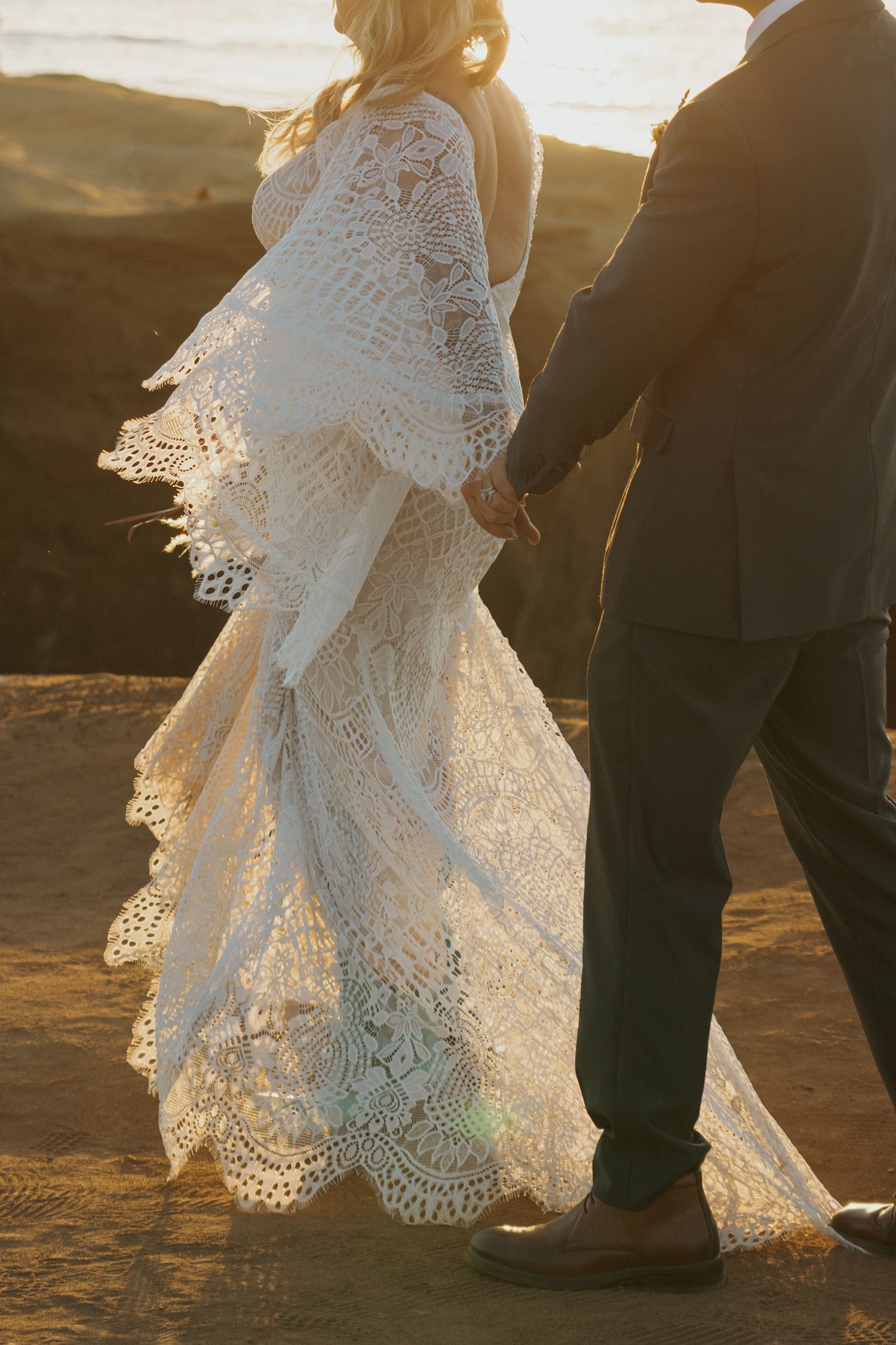 Elegant lace wedding dress flowing in sunset light as couple stands on cliffside.