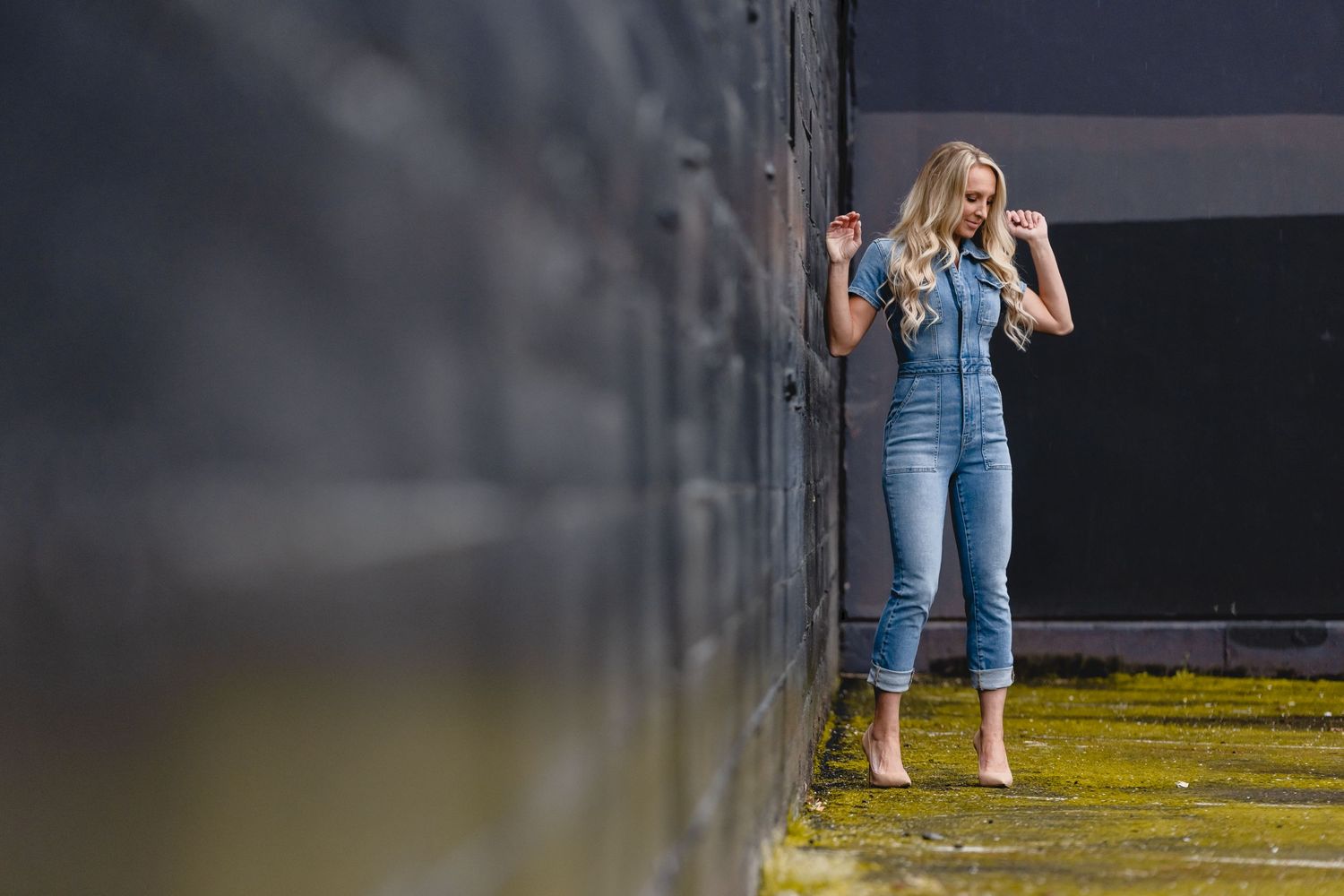 A person in denim overalls and yellow boots posing against a wall in an artistic setting.