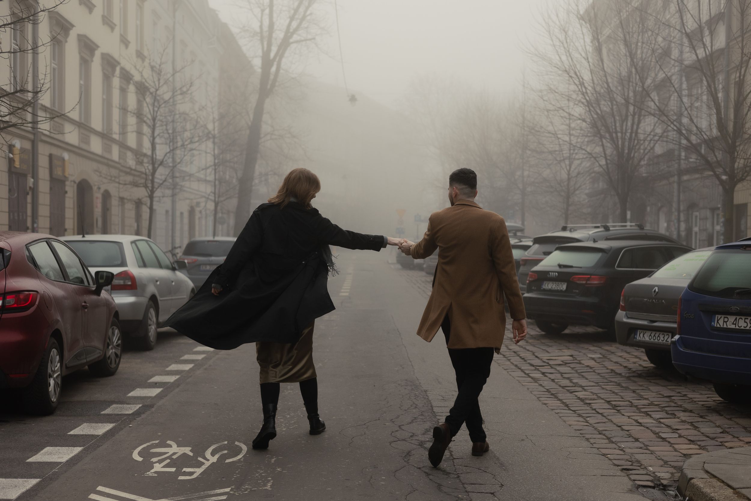 Two people in winter coats walk together on a foggy city street lined with parked cars and buildings.