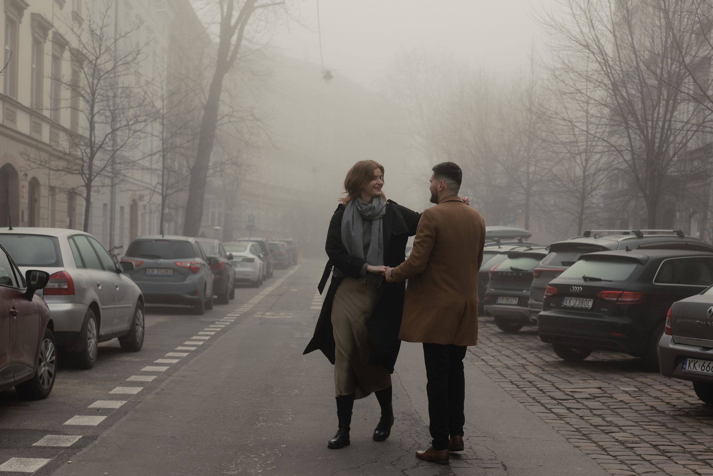 Two people walking together and conversing on a misty city street lined with parked cars on a foggy winter day.