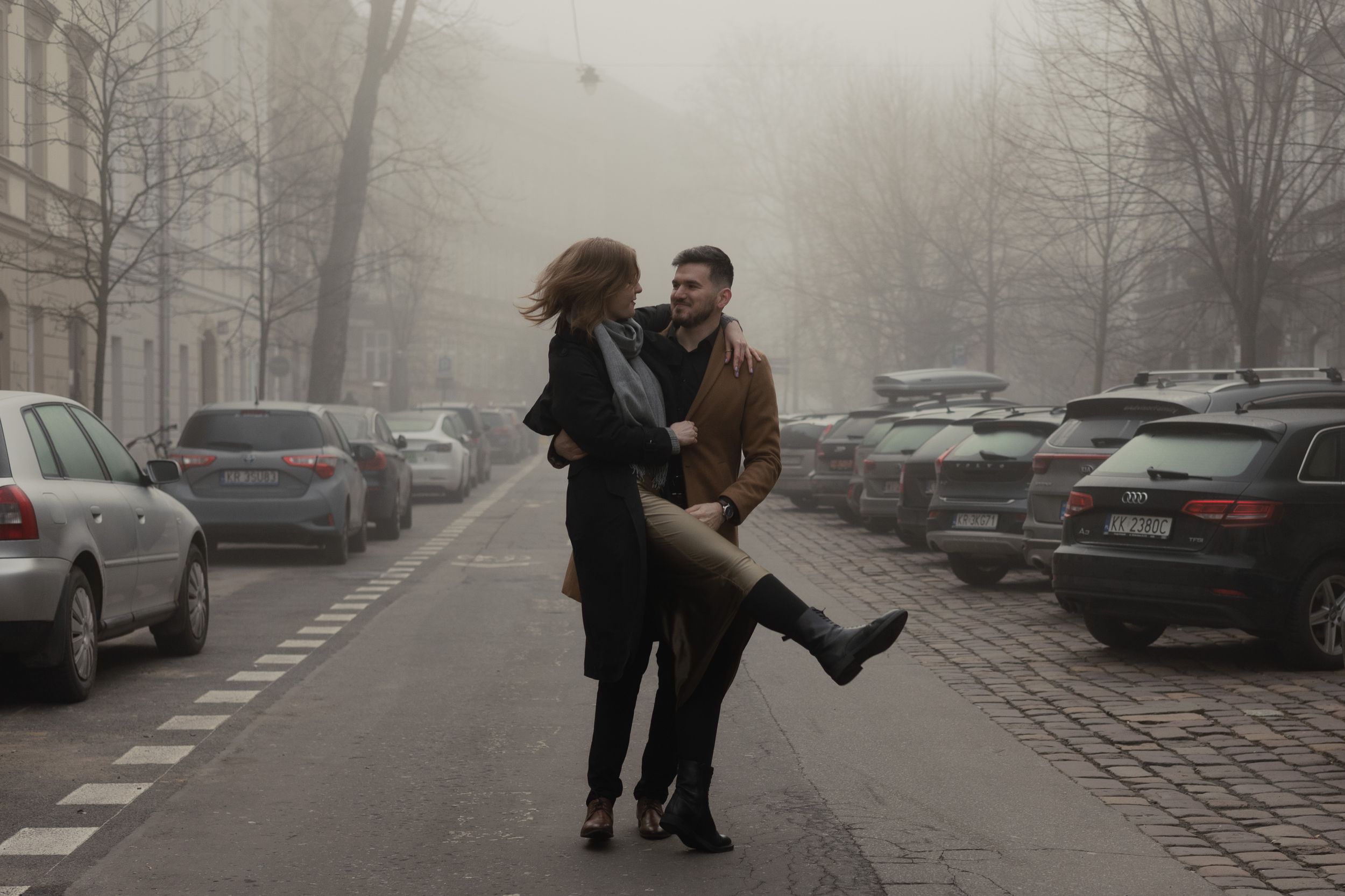 A couple shares a romantic dance moment in a foggy parking lot surrounded by parked cars on a misty day.
