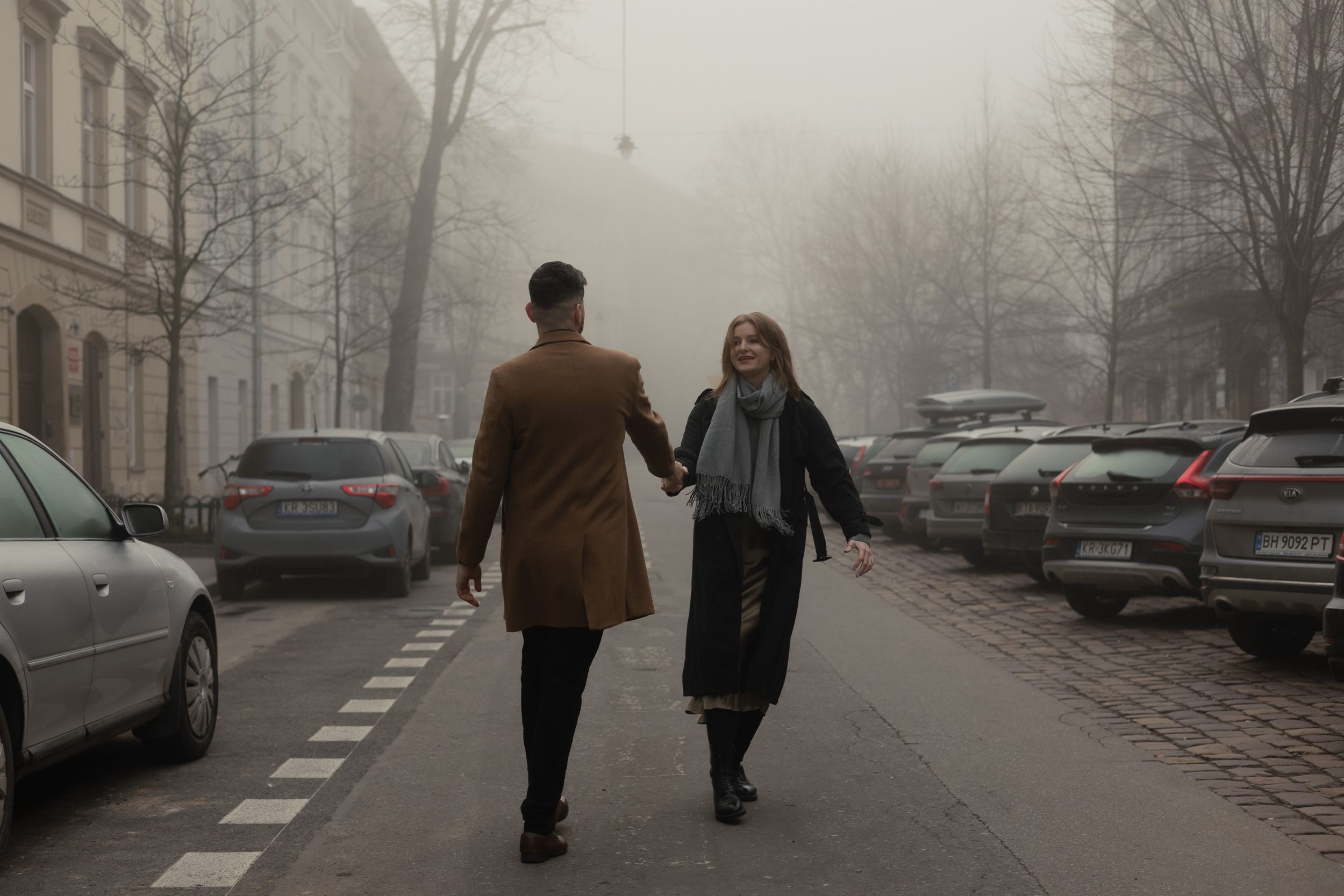 Two people walk together holding hands on a misty winter street lined with parked cars.