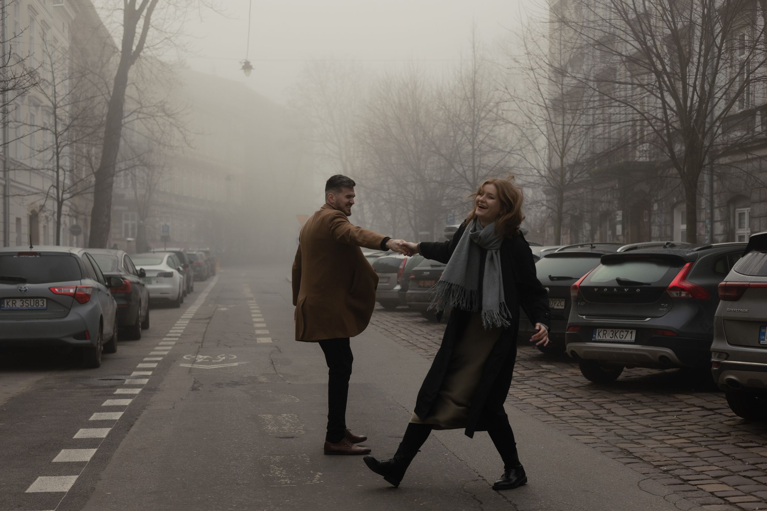 Two people greet each other on a foggy winter street lined with parked cars.