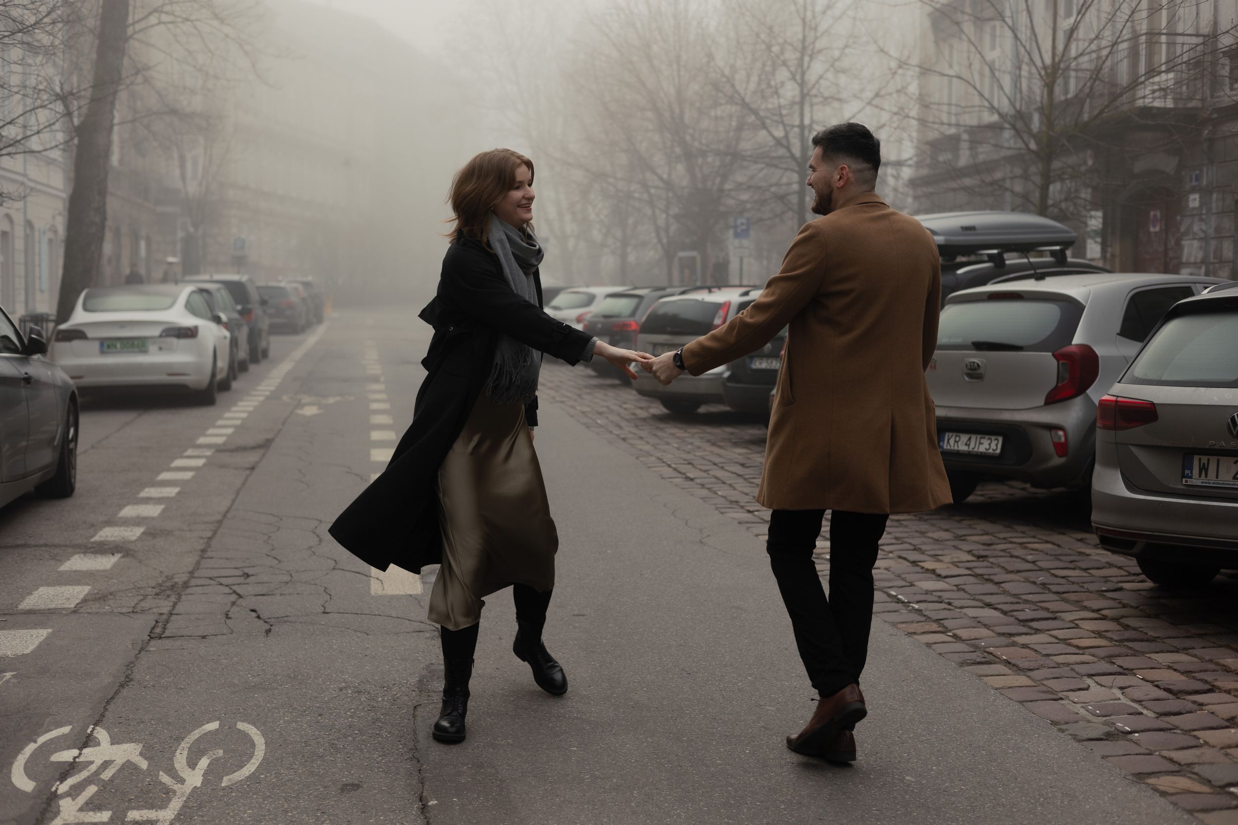 Two people playfully dance together on a foggy street lined with parked cars on a gloomy winter day.