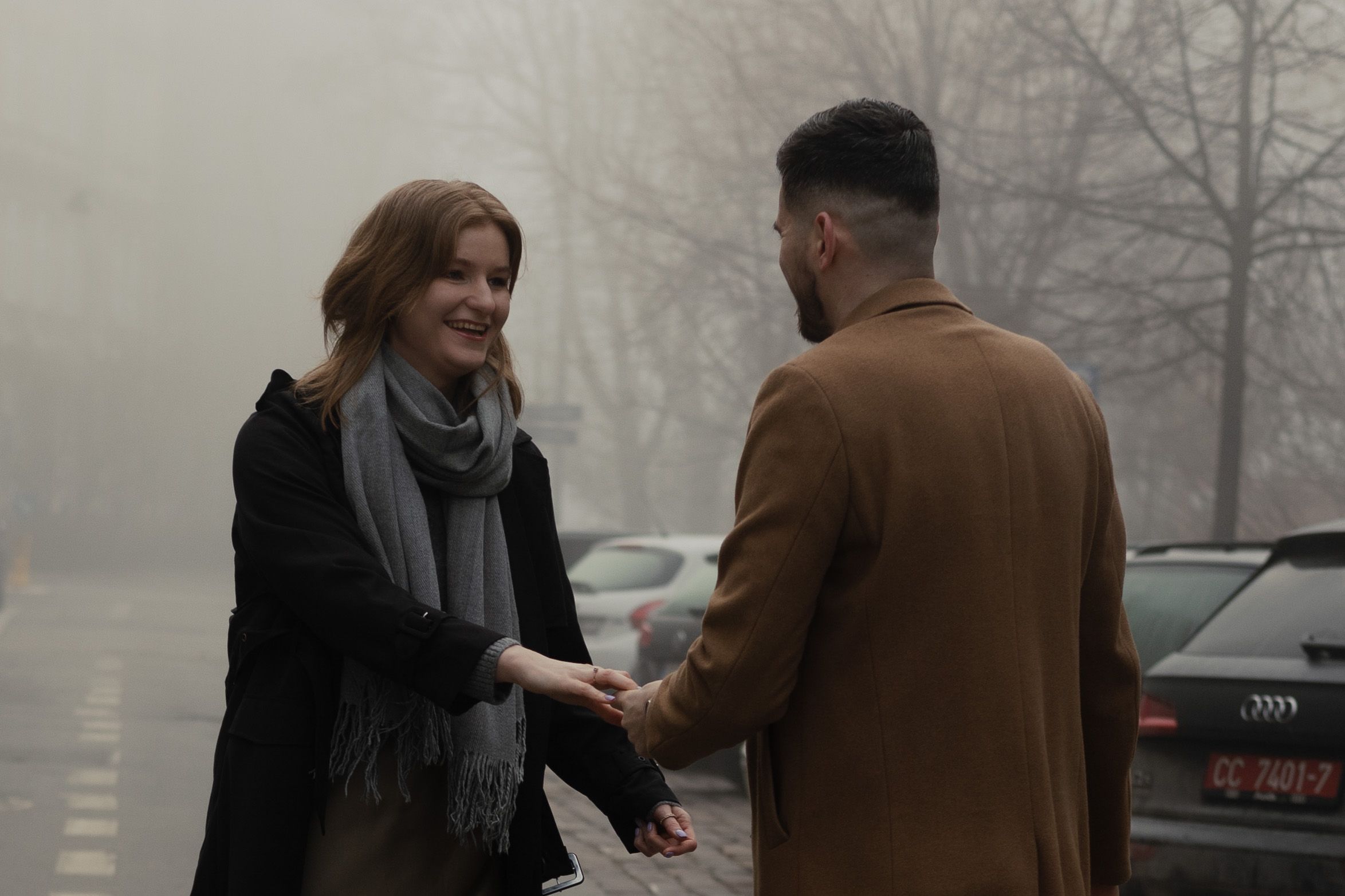 Two people shake hands on a misty winter day in a parking lot wearing warm coats and scarves.