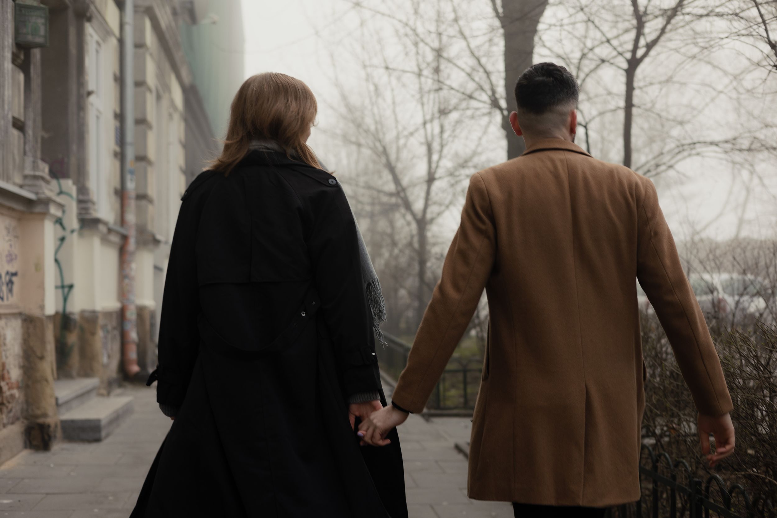 A couple in coats holds hands while walking down a winter sidewalk on a gloomy day.