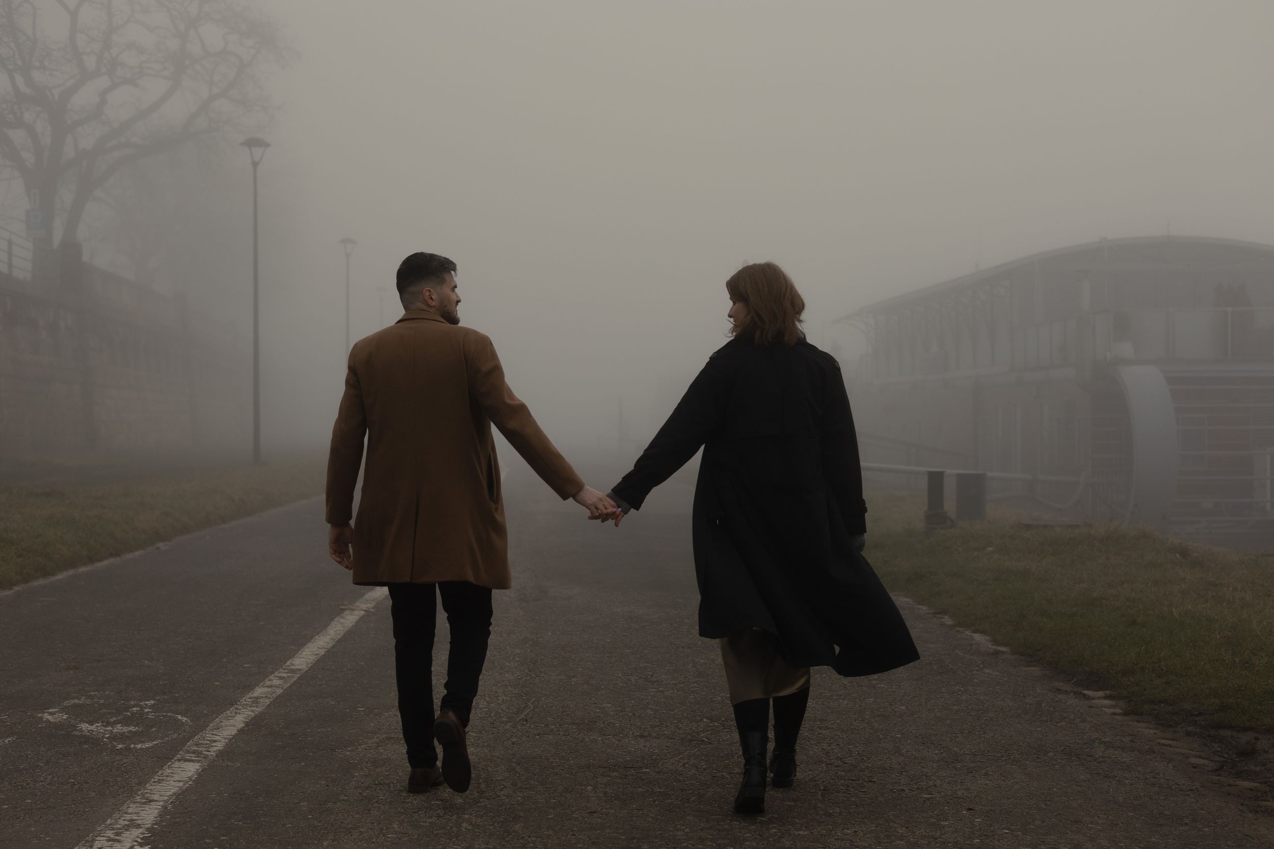 A couple holds hands while walking on a foggy path wearing winter coats in atmospheric misty weather.