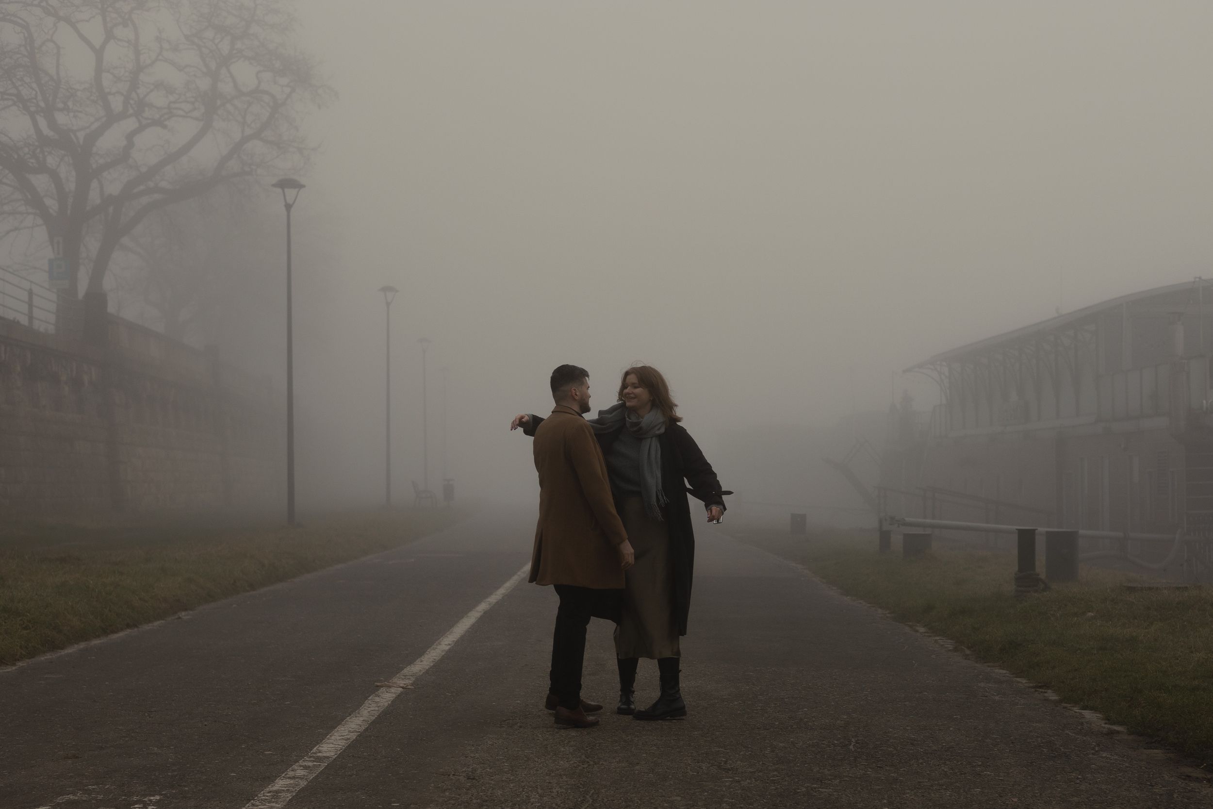Two individuals walk together on a misty path with buildings visible in the foggy background.