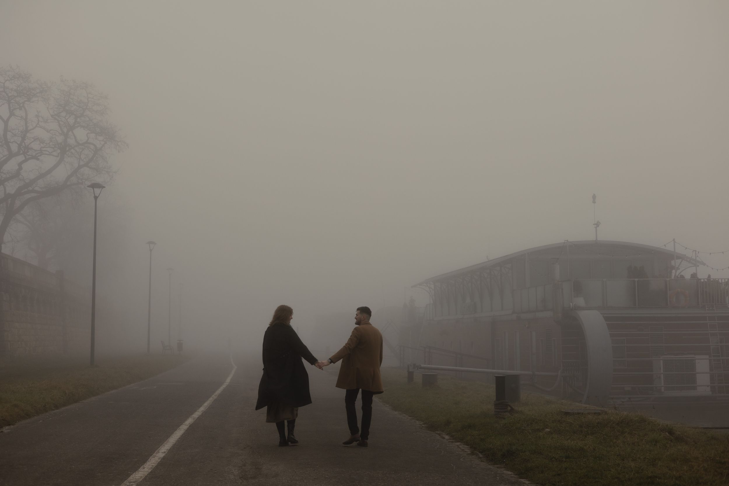 Two figures walk hand in hand along a foggy road beside an abandoned building on a misty day.