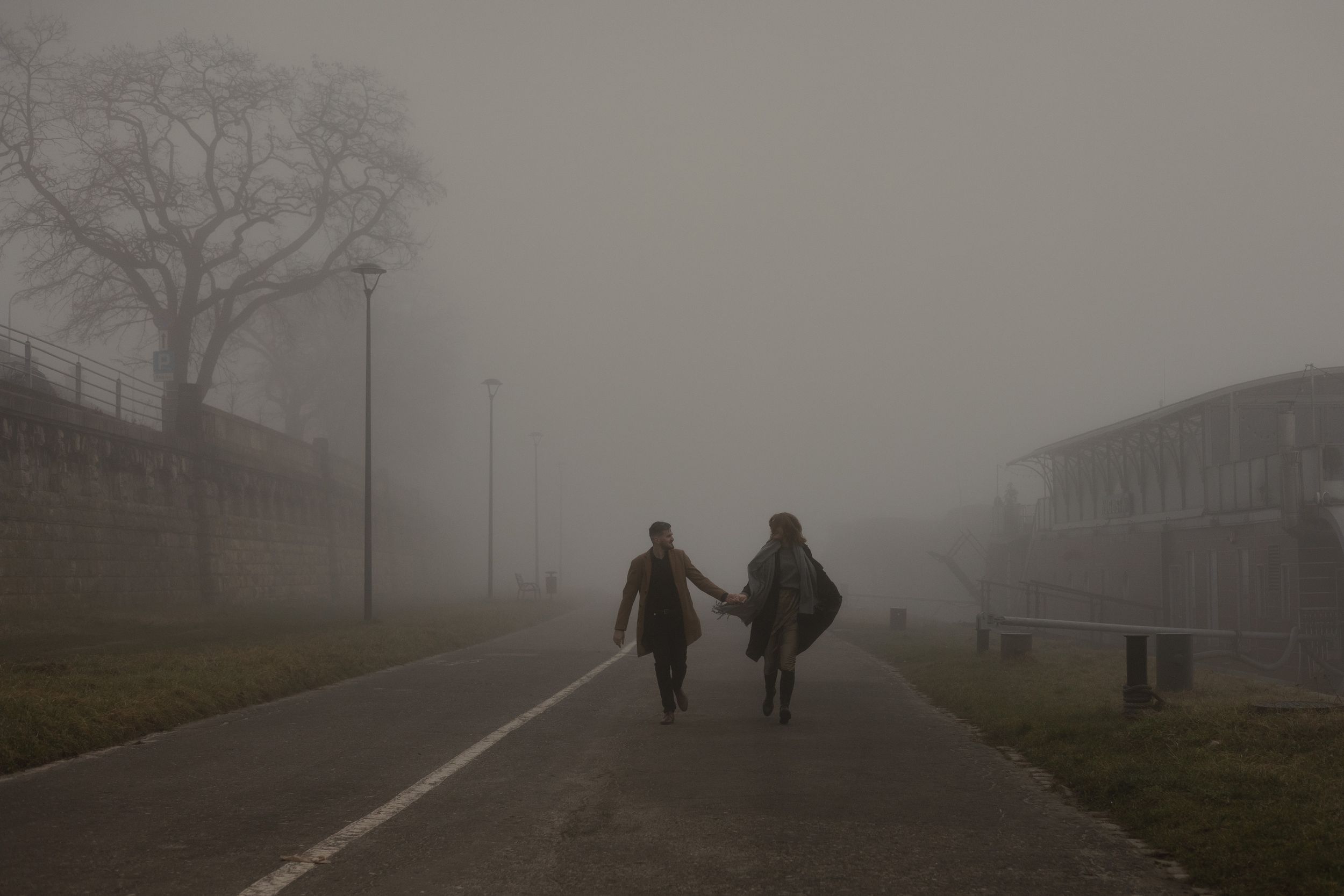 Silhouettes of two people walking away on a foggy path bordered by grass and buildings.