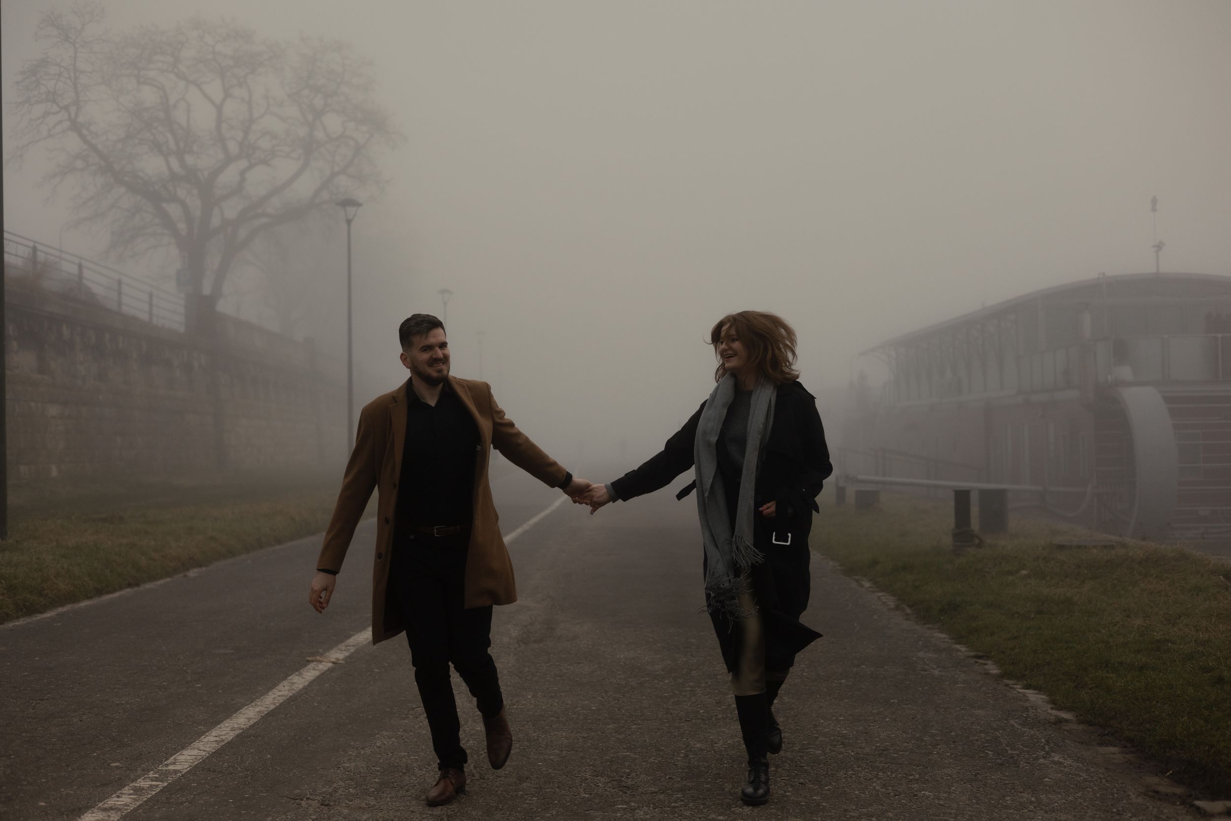 Two people hold hands while walking together on a misty road with buildings in the background.