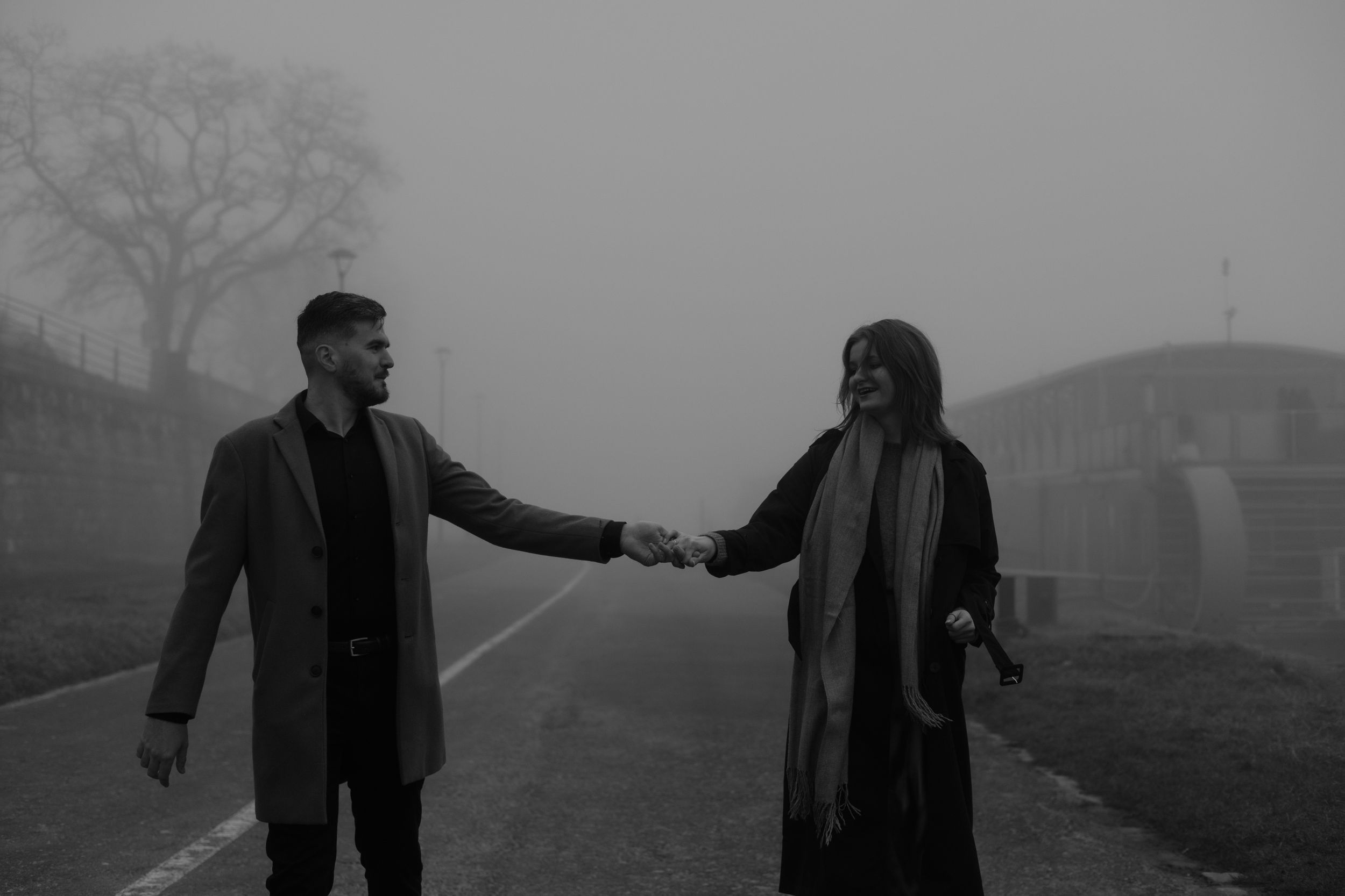 Two people in dark coats hold hands while standing on a misty road in black and white.