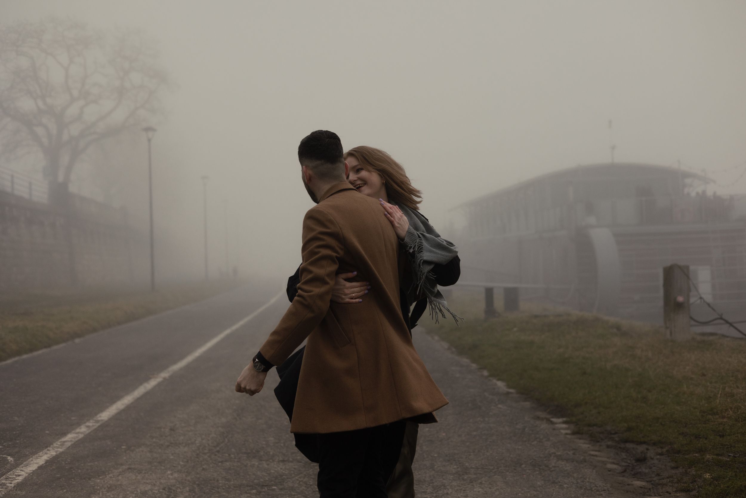 Two people share an intimate embrace on a foggy street with a misty background and houses visible in the distance.