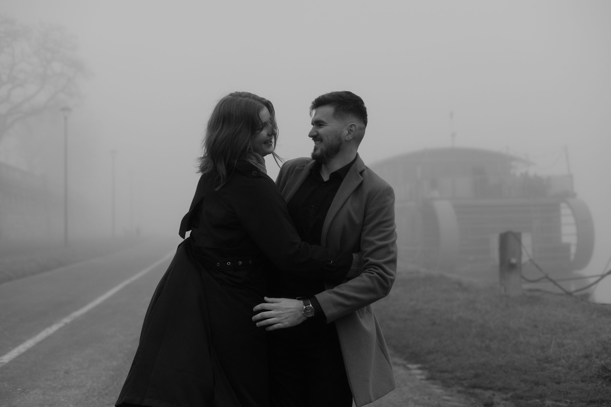 A couple in winter coats embrace on a foggy day in black and white photography.