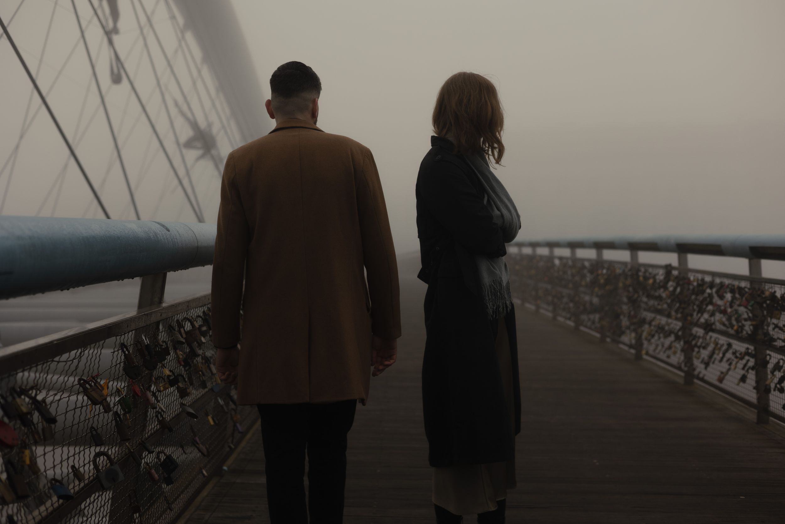 Two people in coats stand together on a misty wooden bridge during a foggy day.