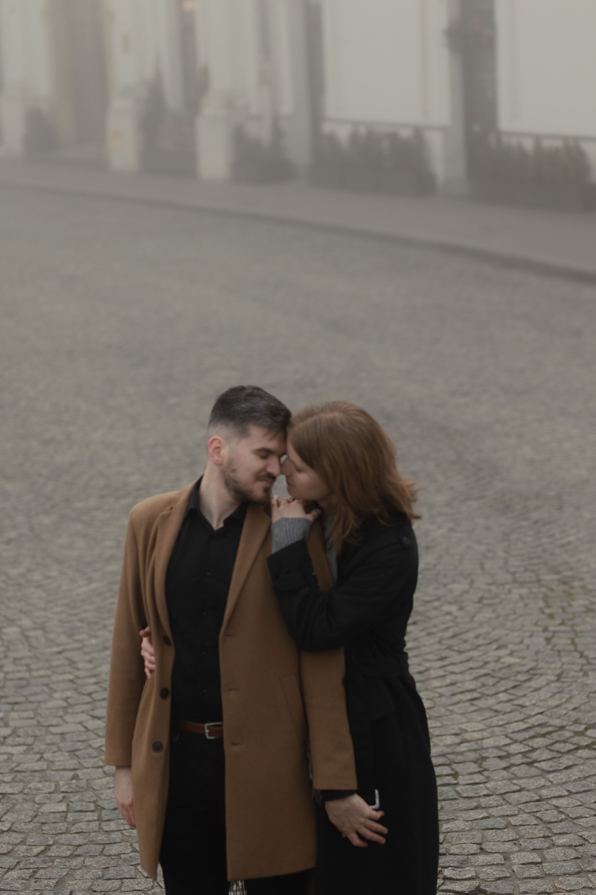 A romantic couple in warm coats sharing an intimate moment on a foggy cobblestone street.