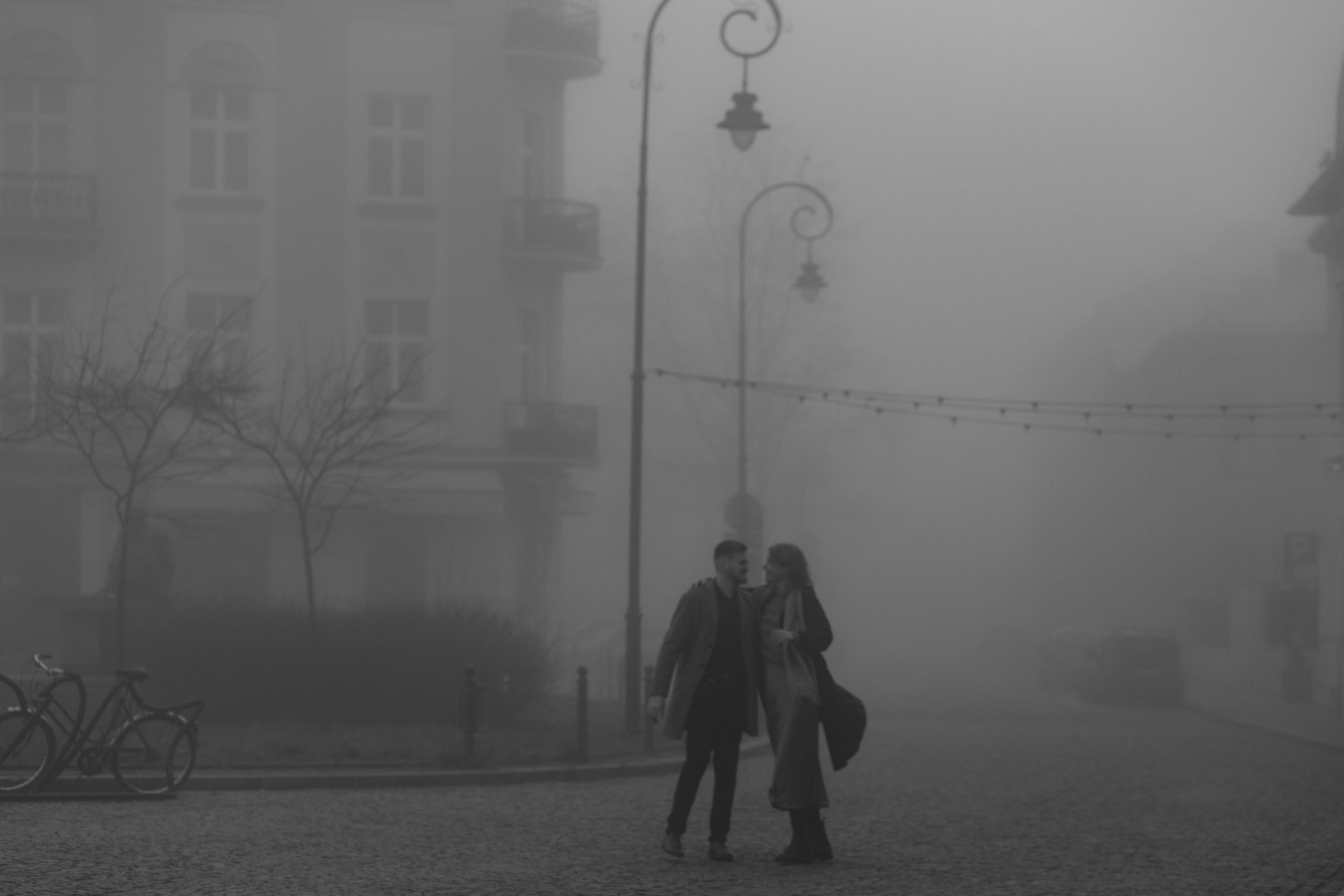Silhouettes of people walking together on a foggy city street with ornate lampposts and buildings in the background.