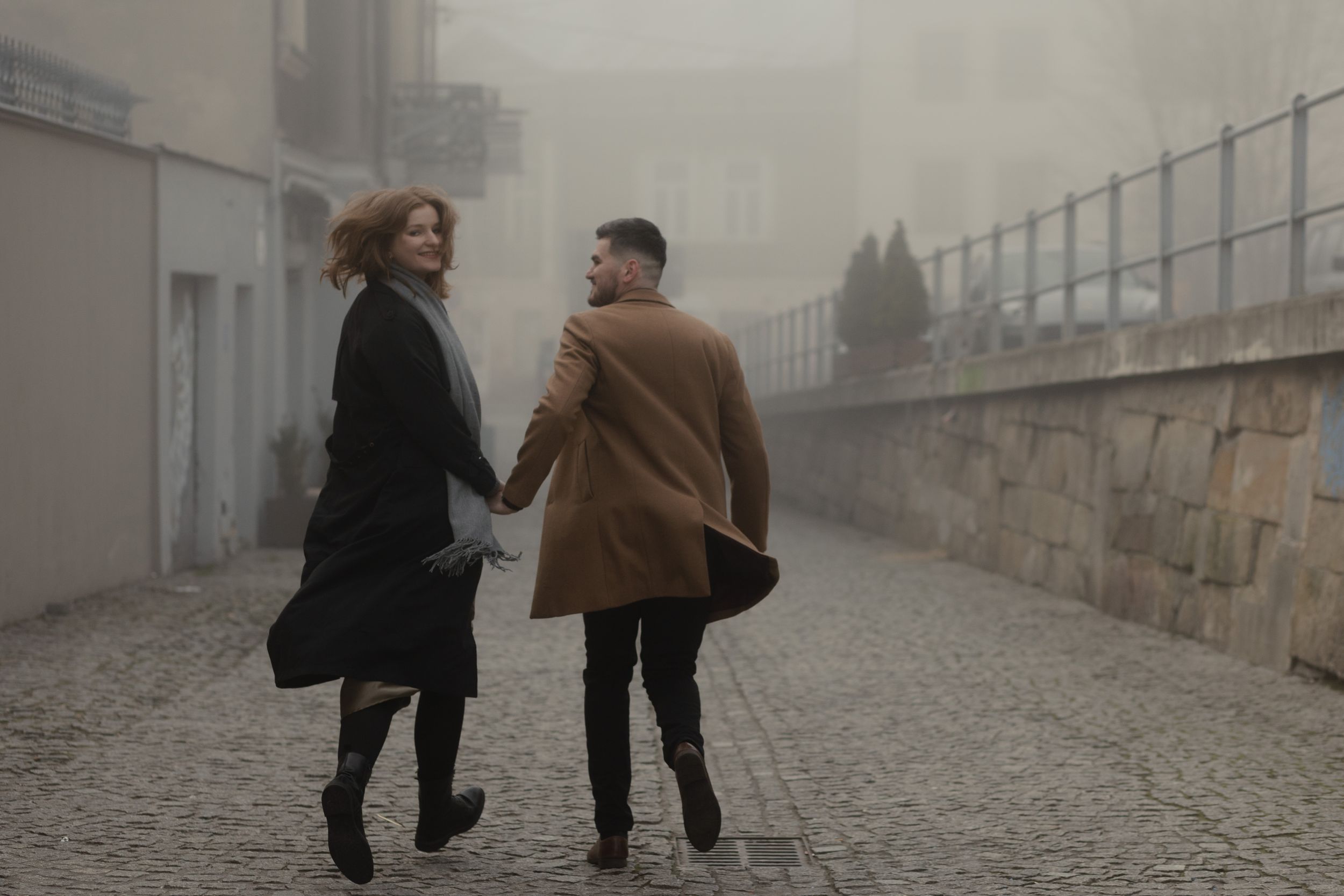 Two people walking and holding hands on a foggy cobblestone path beside a stone wall.