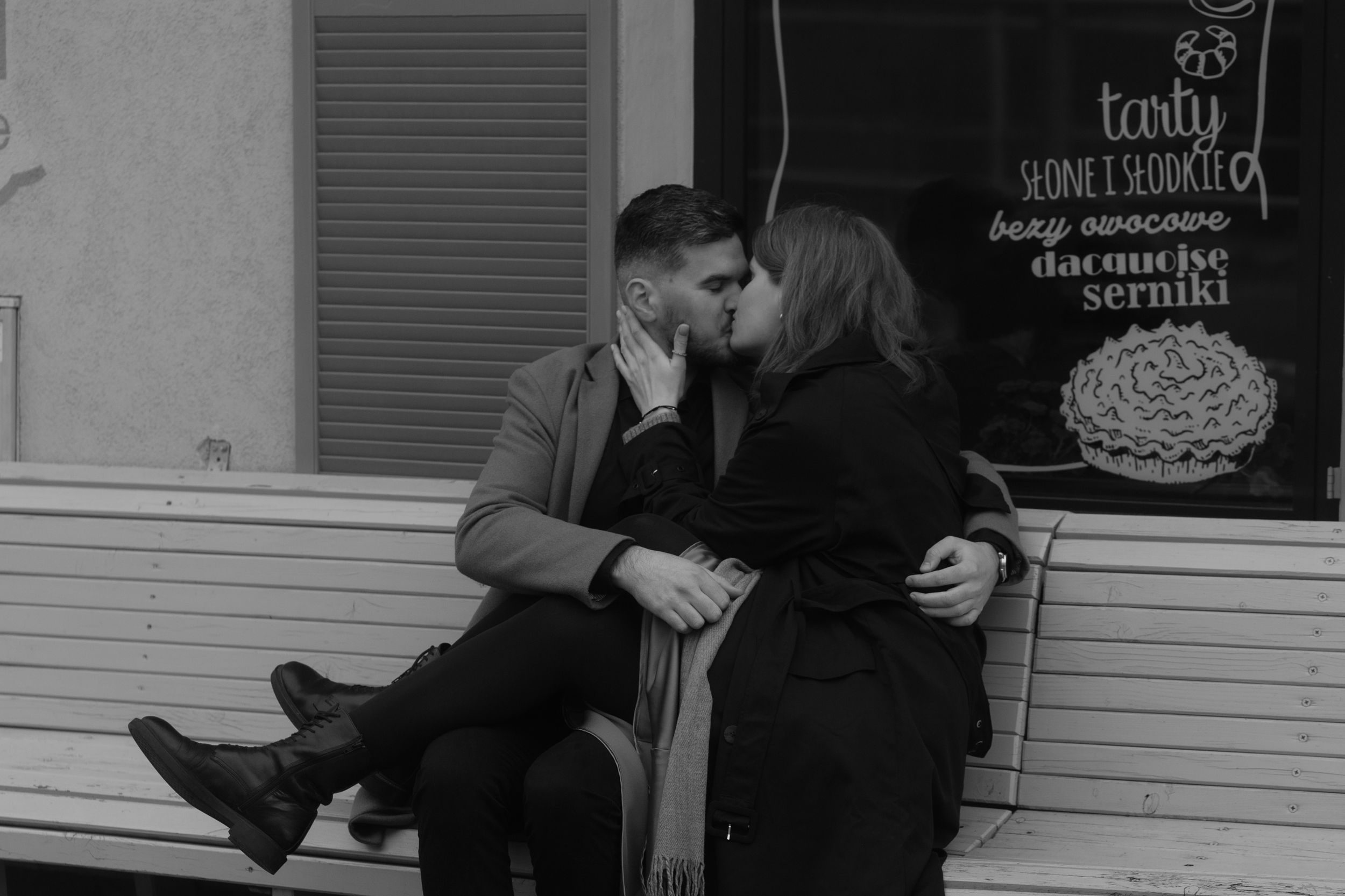 A romantic black and white photo of a couple sitting close together on a bench outside a storefront.