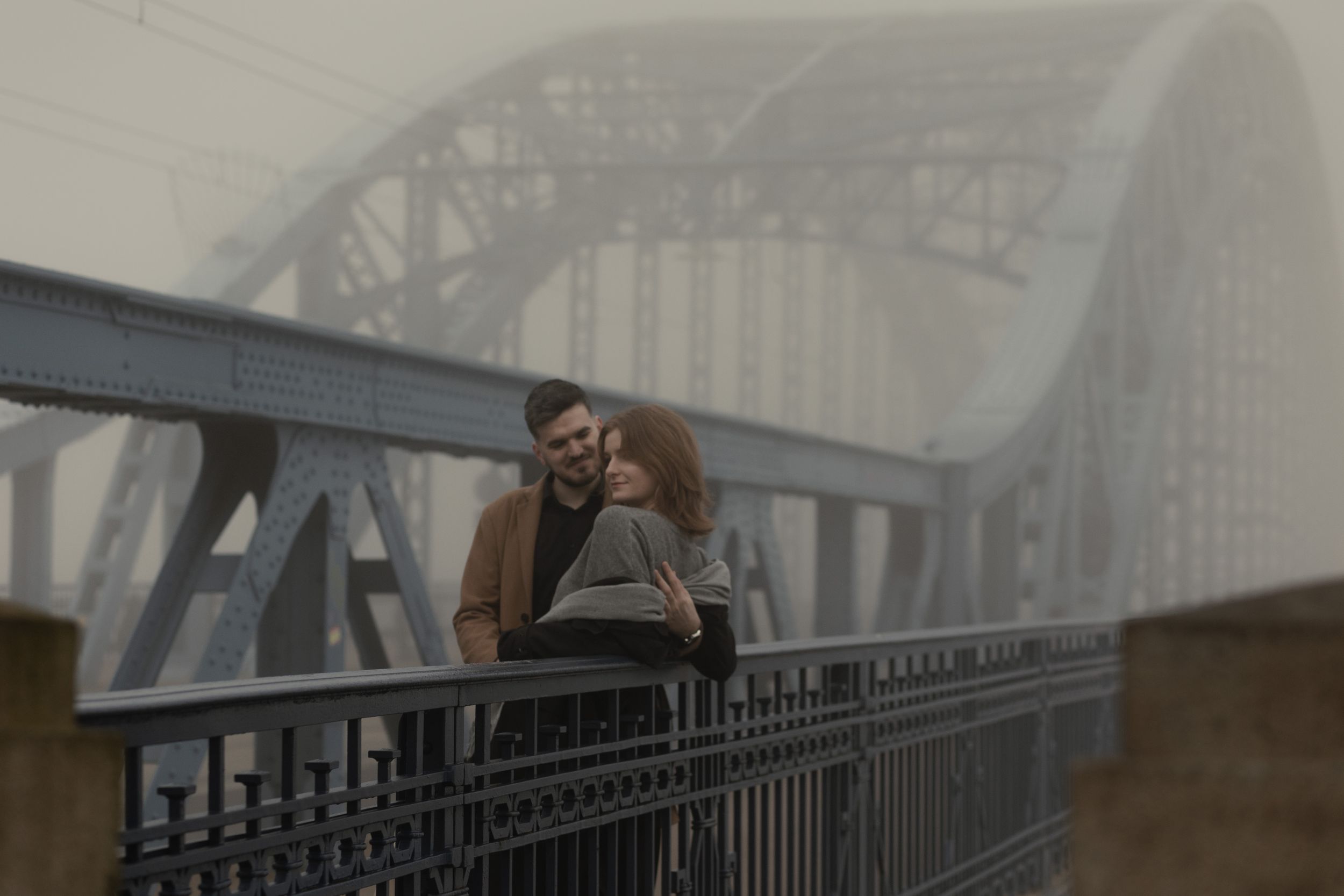 A couple embraces on a foggy metal bridge, creating a moody atmospheric scene.