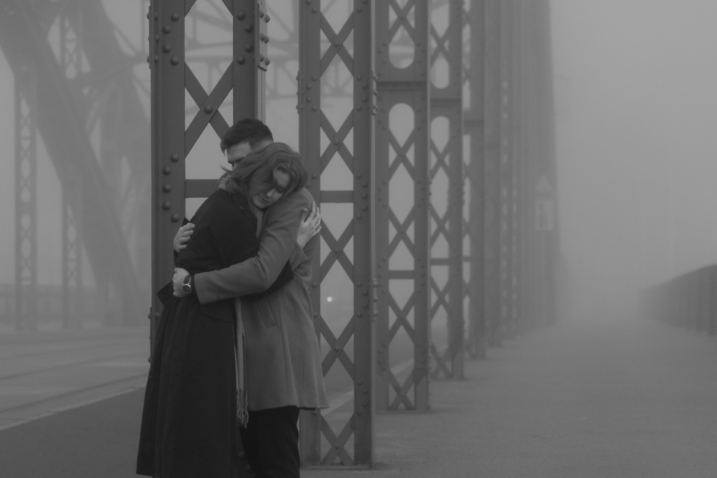 A couple embraces in the foggy mist on a bridge with metal beams and architectural elements visible in the background.