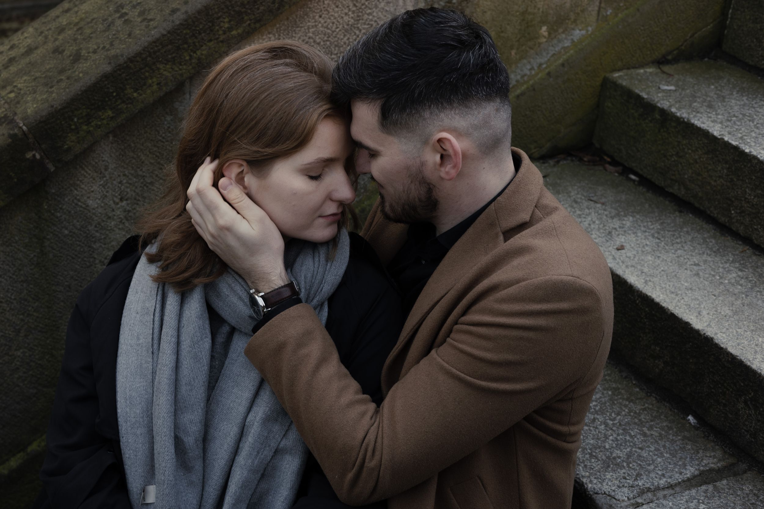 Two people share an intimate moment on stone steps as they embrace, with one wearing a brown coat and the other in grey.
