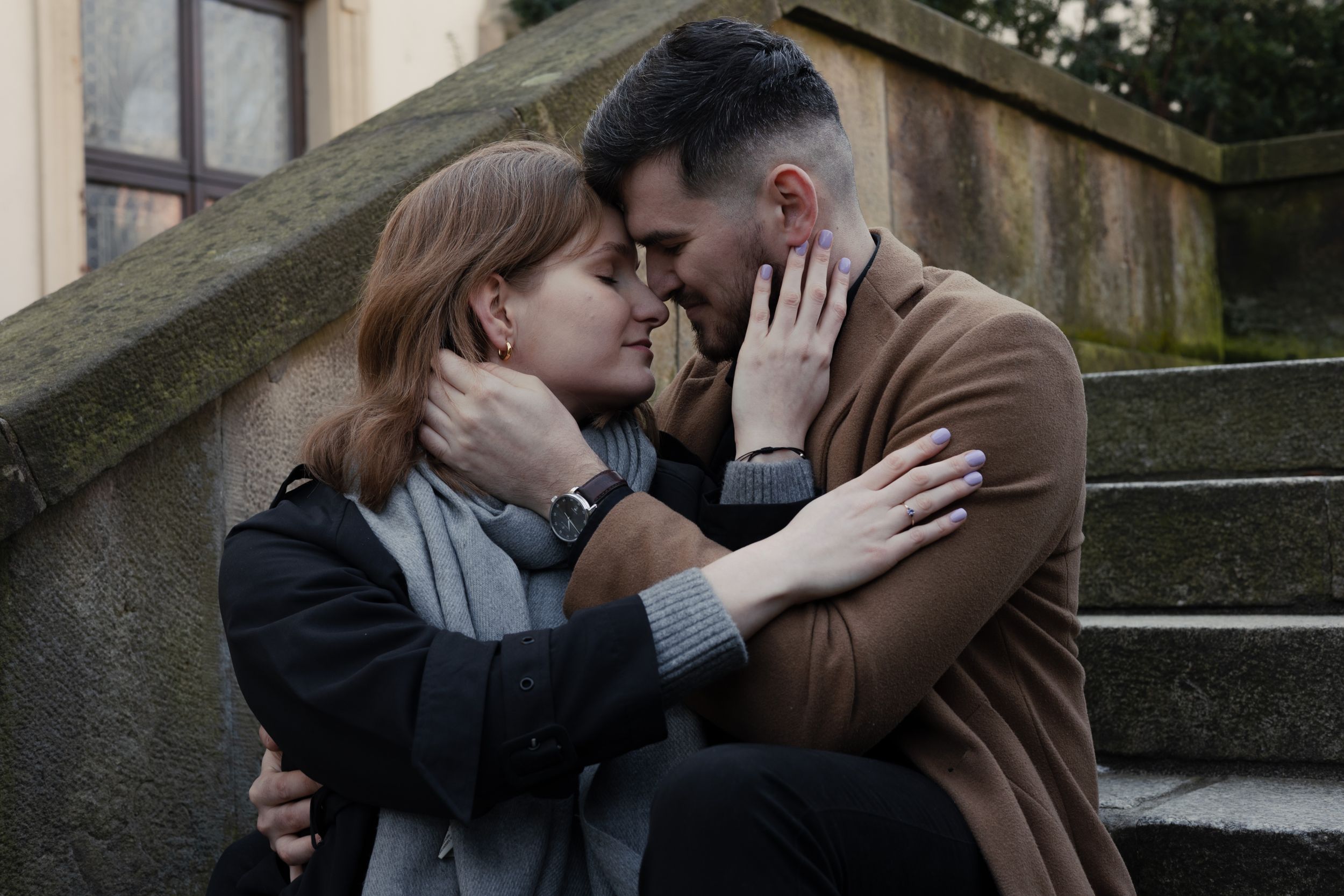 A tender romantic moment between two people embracing on stone steps, wearing winter coats on an overcast day.