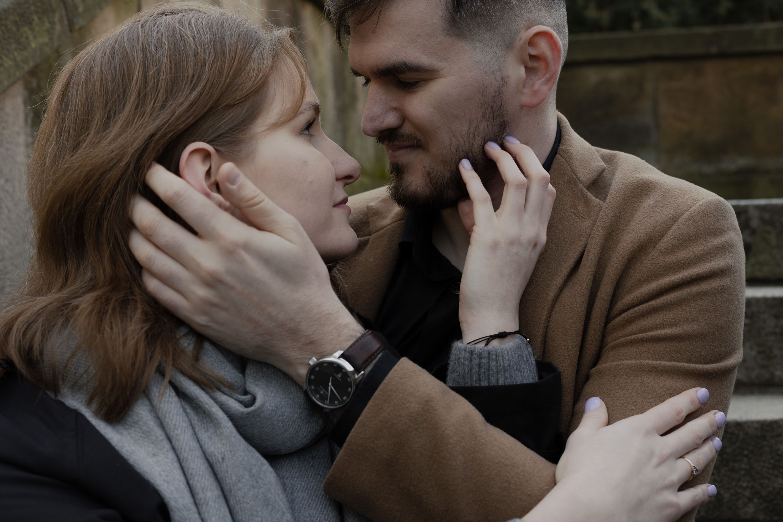 Two people share an intimate moment outdoors as they lovingly embrace and touch each others faces with hands.