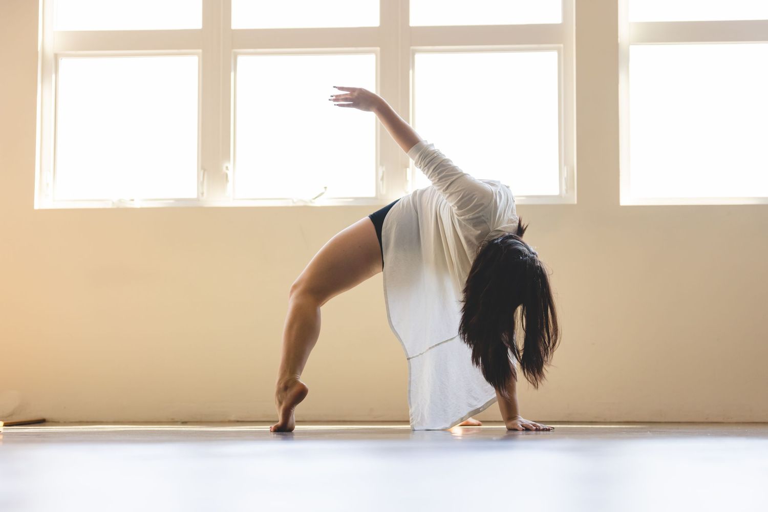 Contemporary dancer in white leotard performs graceful backbend pose in bright sunlit studio space.