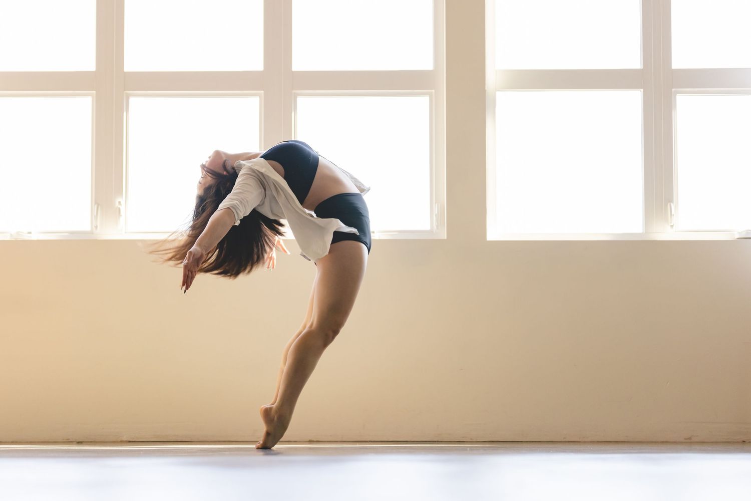 A dancer performs a graceful backbend leap in a sunlit dance studio with large windows.