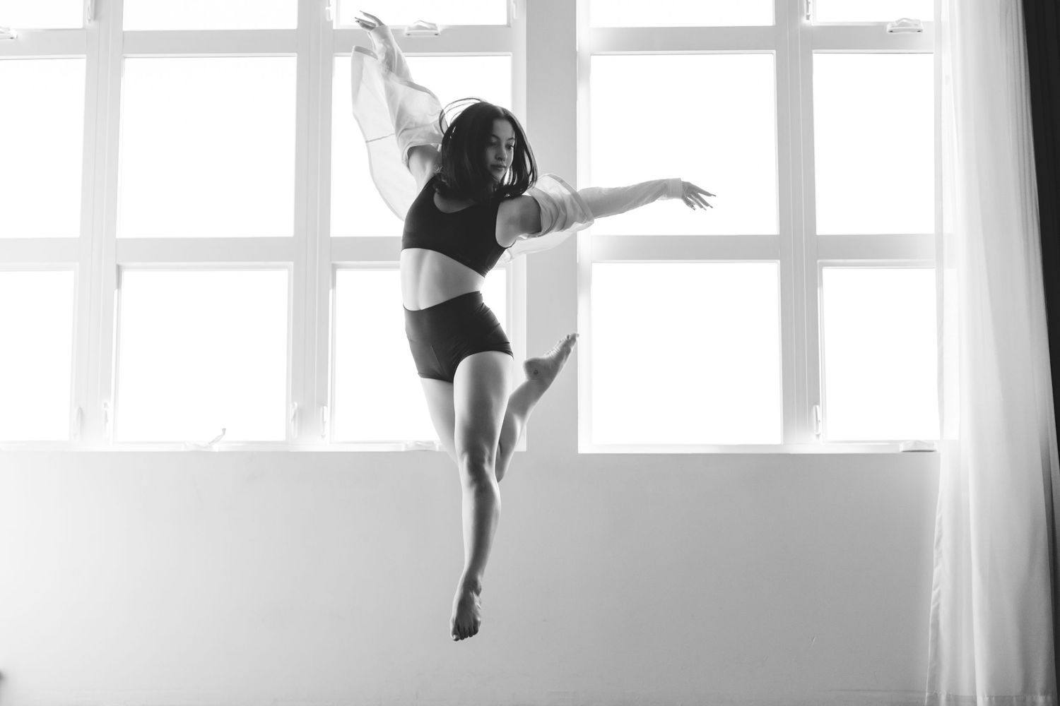 A dancer leaps gracefully in a minimalist studio space with bright backlit windows creating a dramatic silhouette effect.