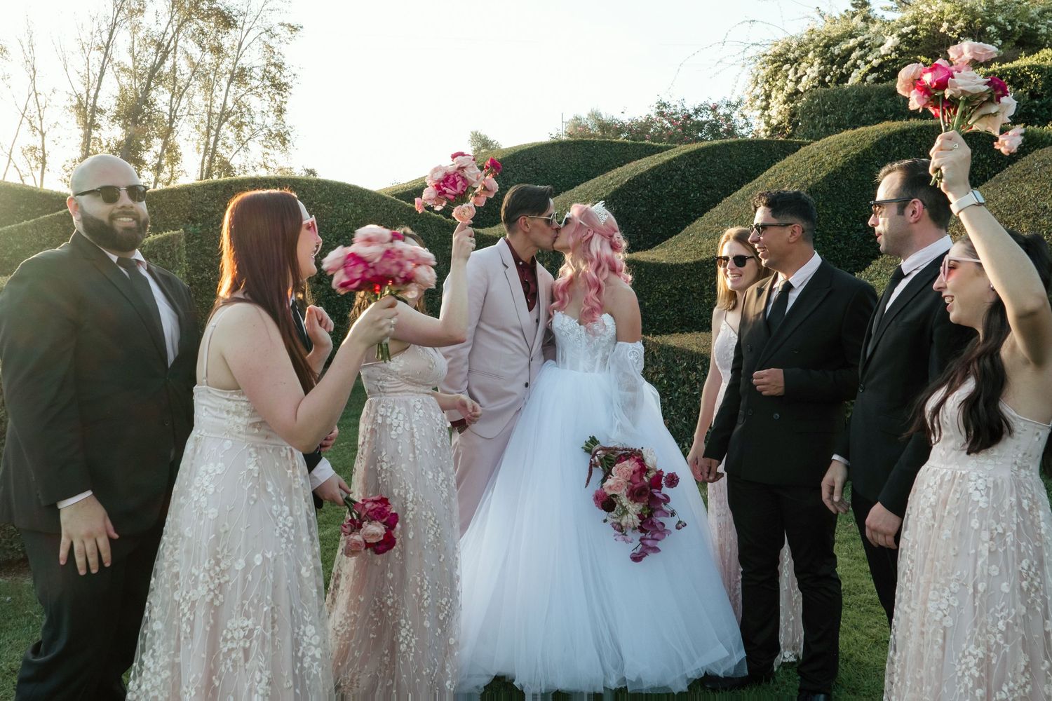 Wedding party celebrates outdoors with bridesmaids in blush dresses and groomsmen in black suits holding pink bouquets.