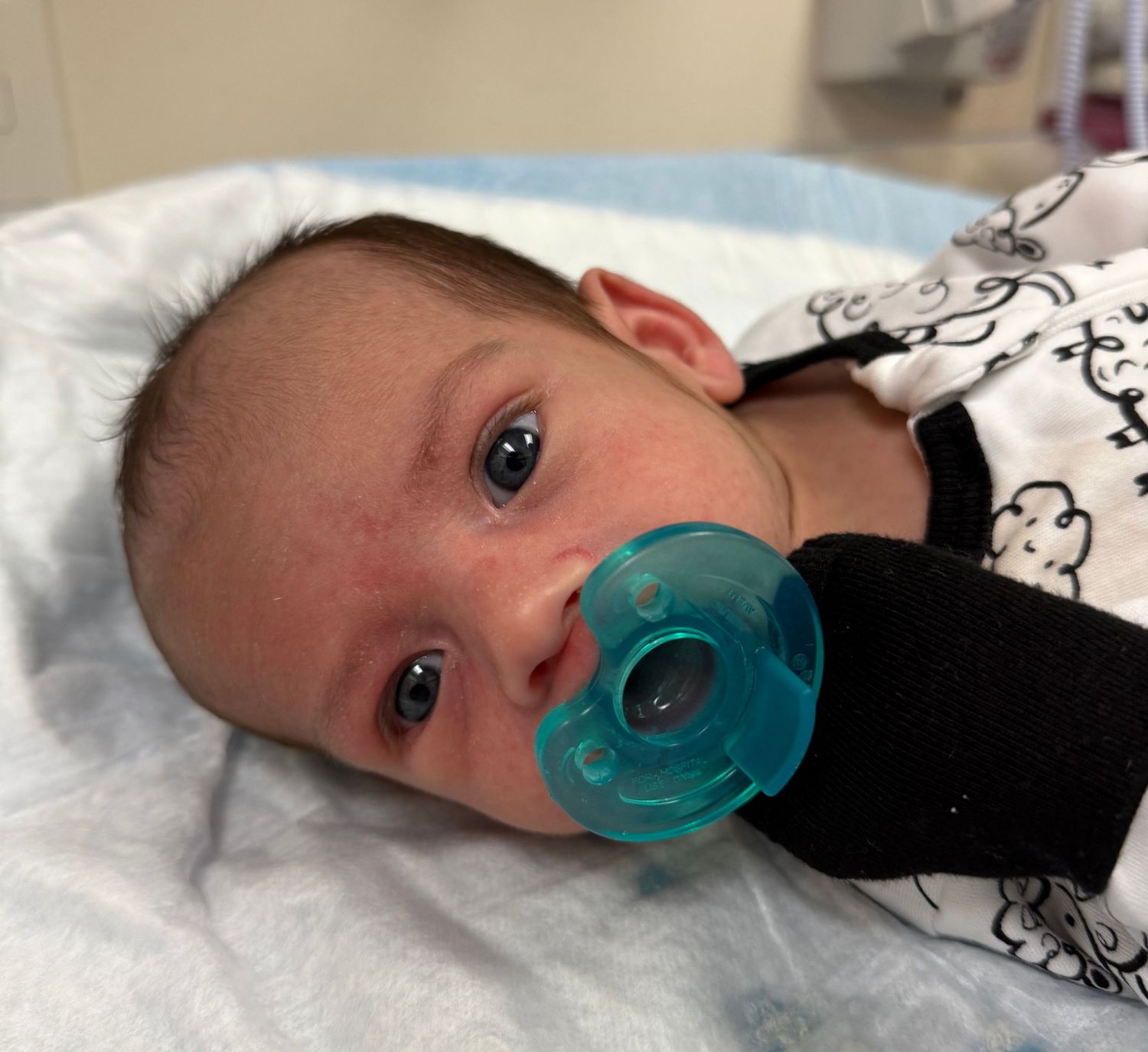 An infant with bright eyes rests on a light blanket while using a teal-colored pacifier.