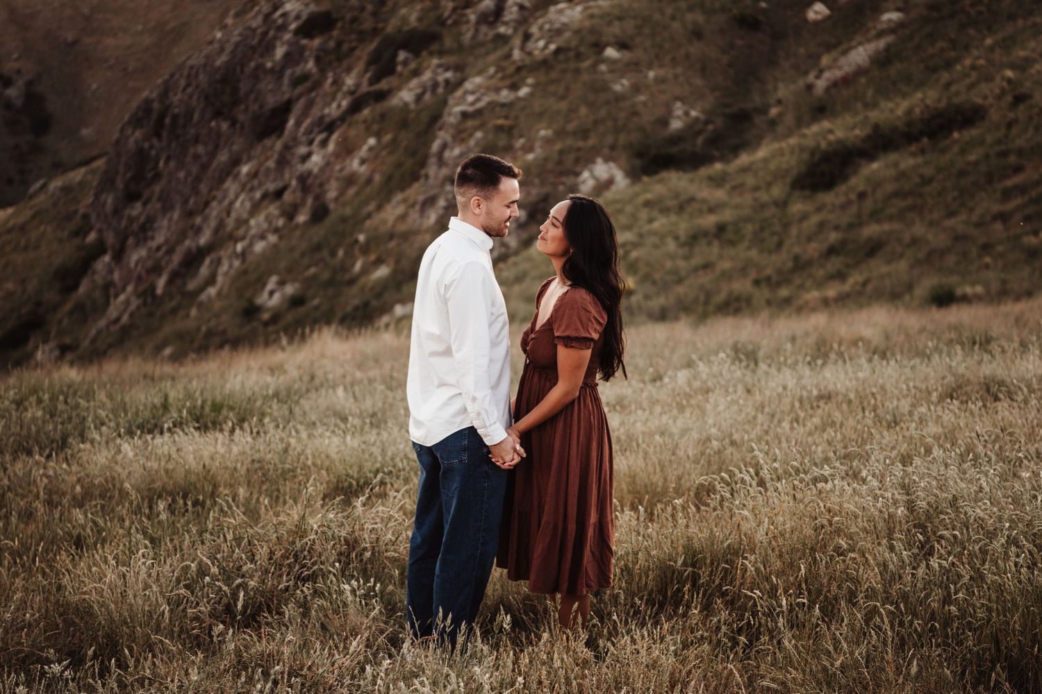 Couple in romantic pose standing in field with mountain backdrop at sunset.