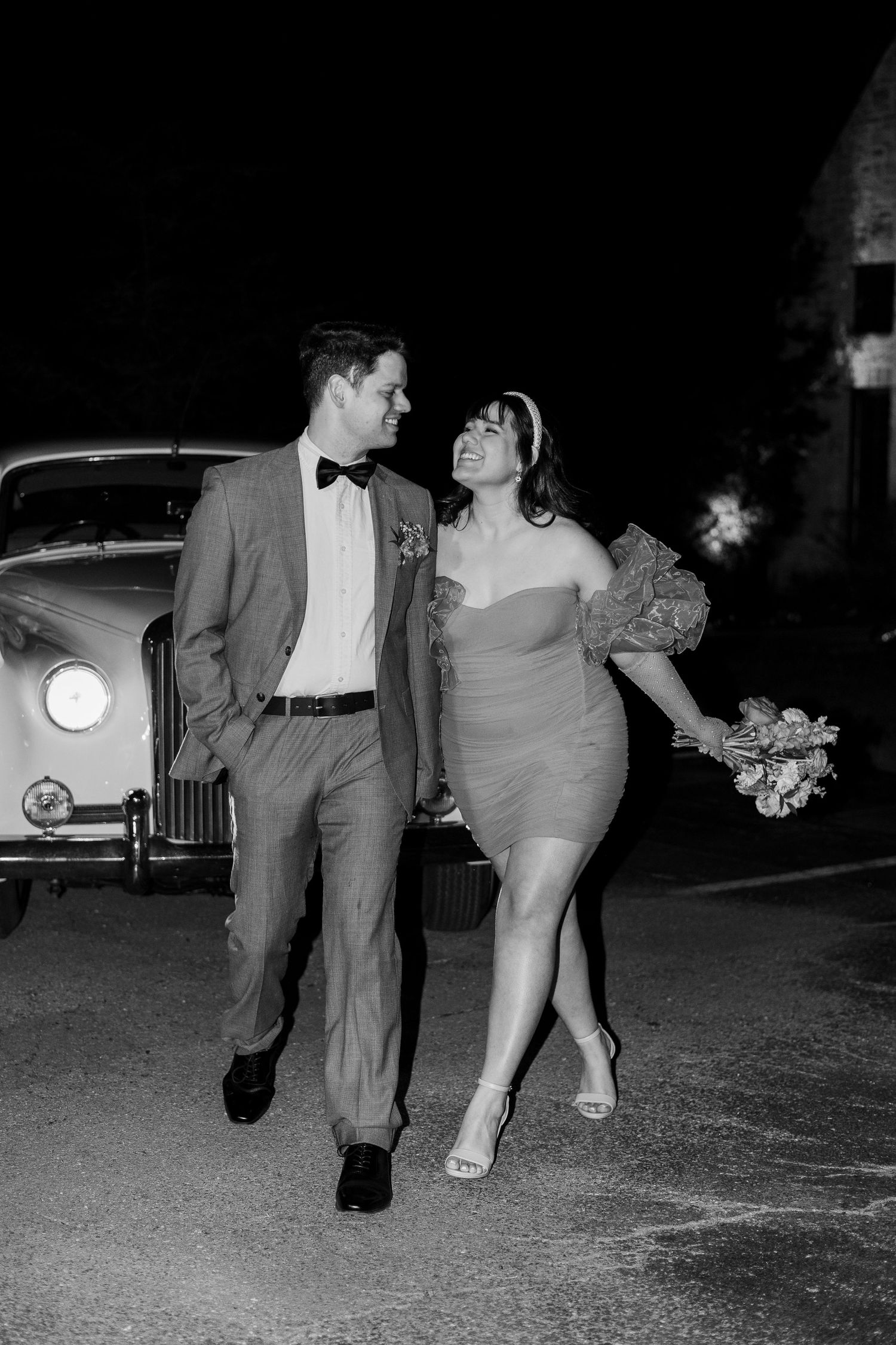 Stylish couple walks past vintage car in black and white nighttime photo.