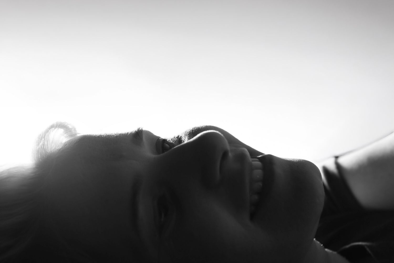 Black and white close-up portrait of a smiling woman lying down with soft backlit glow.