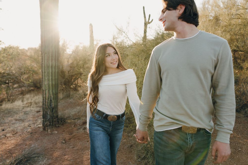 A couple in casual attire walks together through a desert landscape with saguaro cacti at sunset.