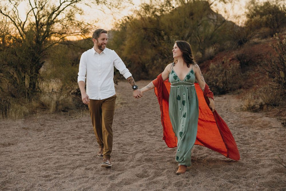 2 People walk hand in hand through a desert landscape during golden hour as a red fabric flows in the breeze in Phoenix Arizona.