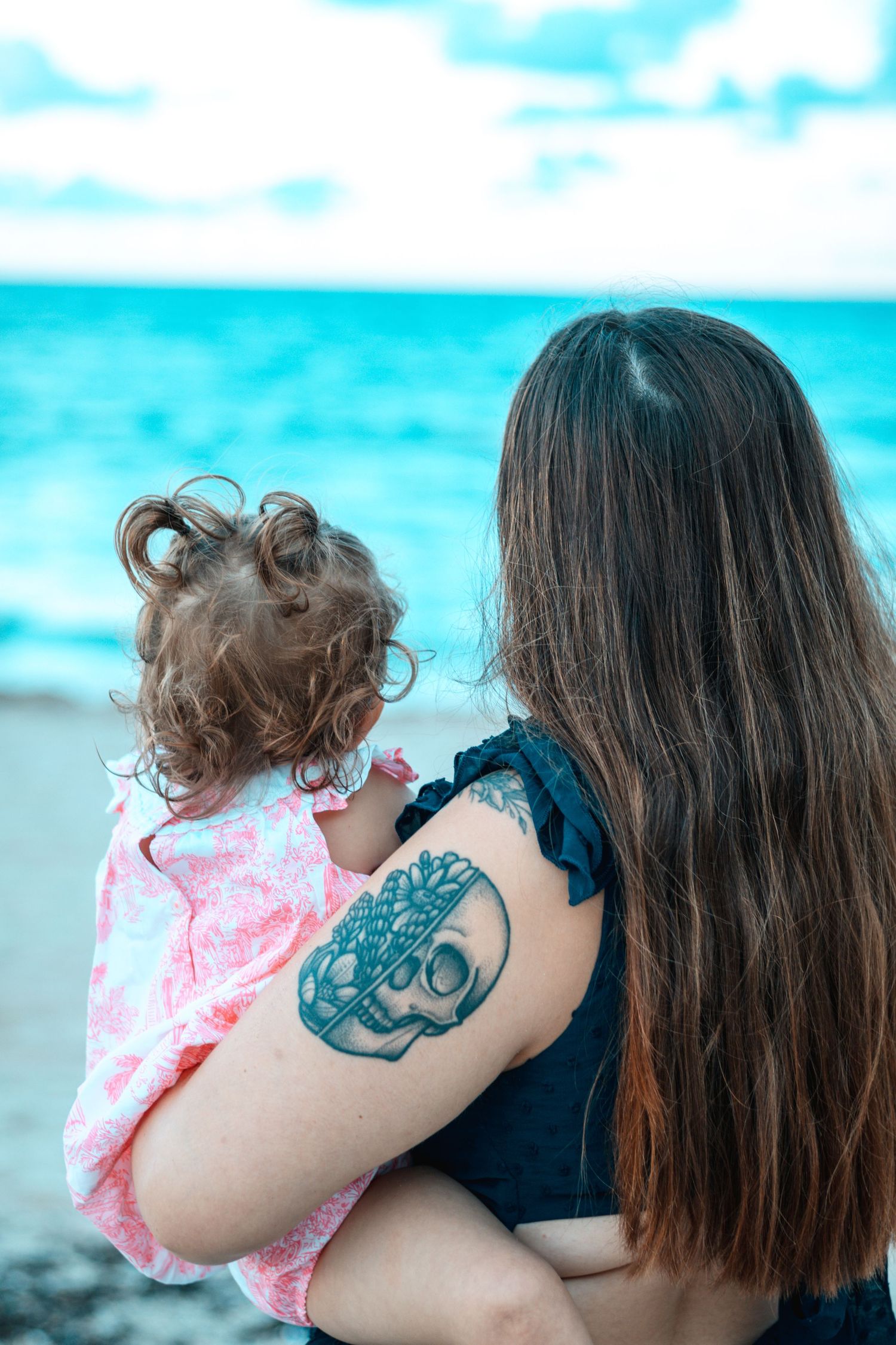 Person with octopus tattoo holding child on beach with blue ocean background.