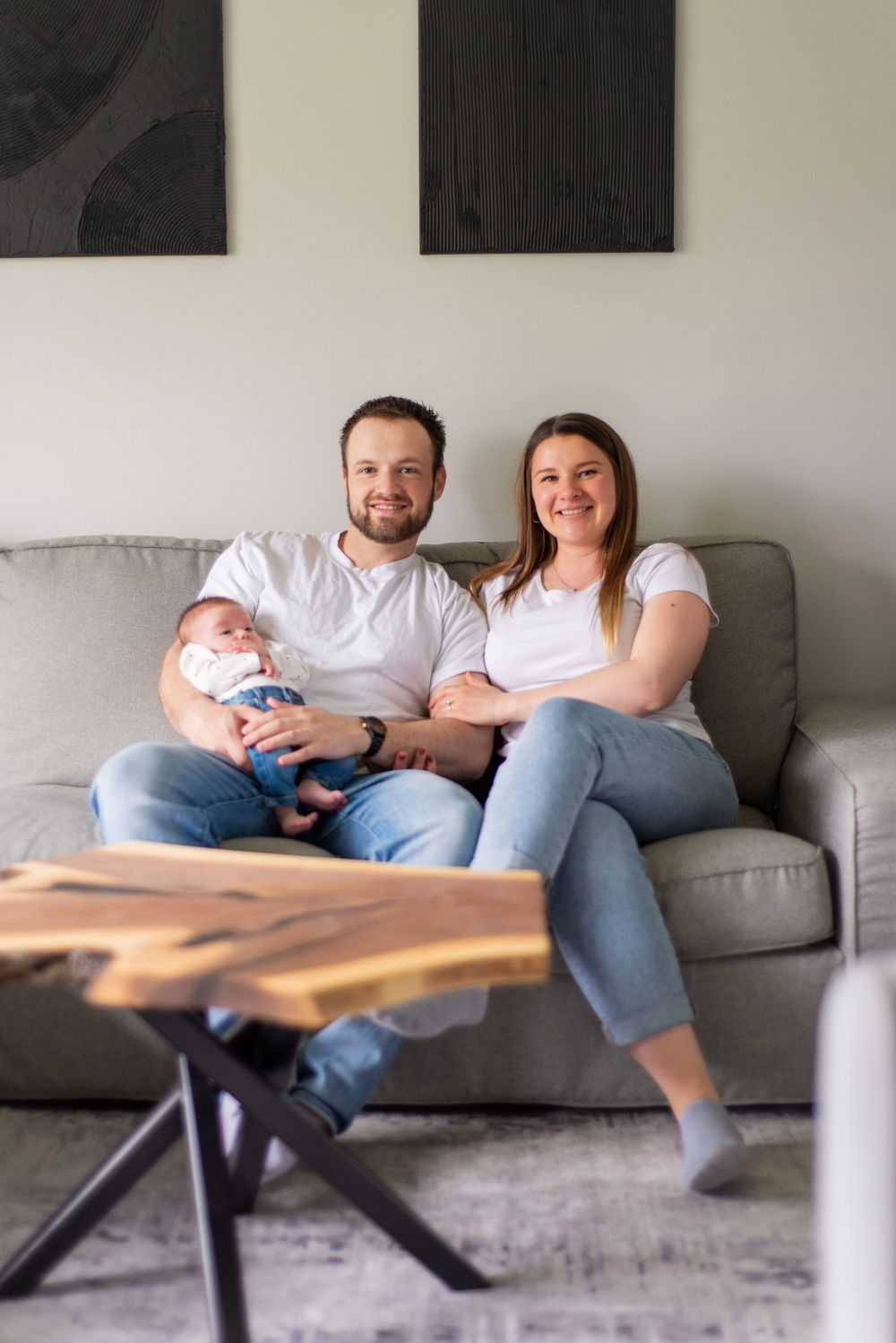 A family portrait on a gray sofa with black artwork hanging on the white wall above.