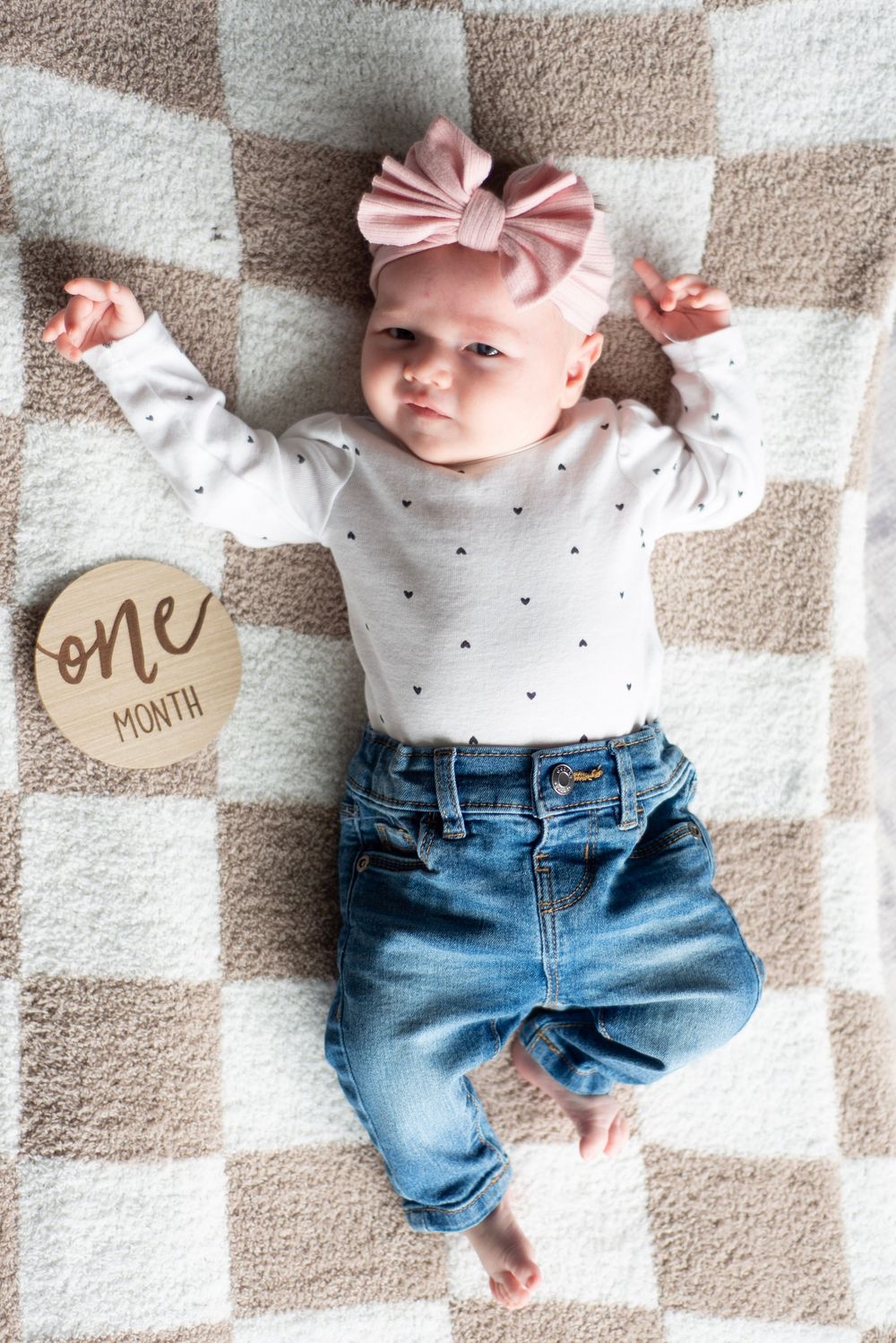 Baby wearing pink bow lies on checkered blanket with 'one month' milestone marker.