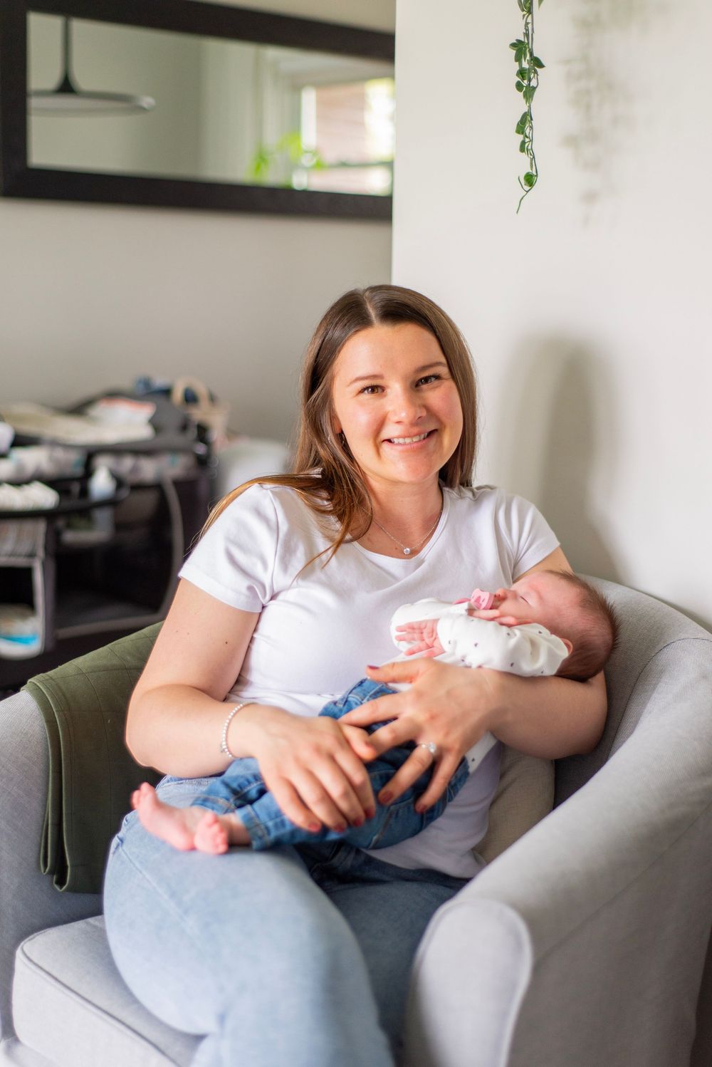 Person sitting in grey armchair holding infant in casual home setting with hanging plant decor.