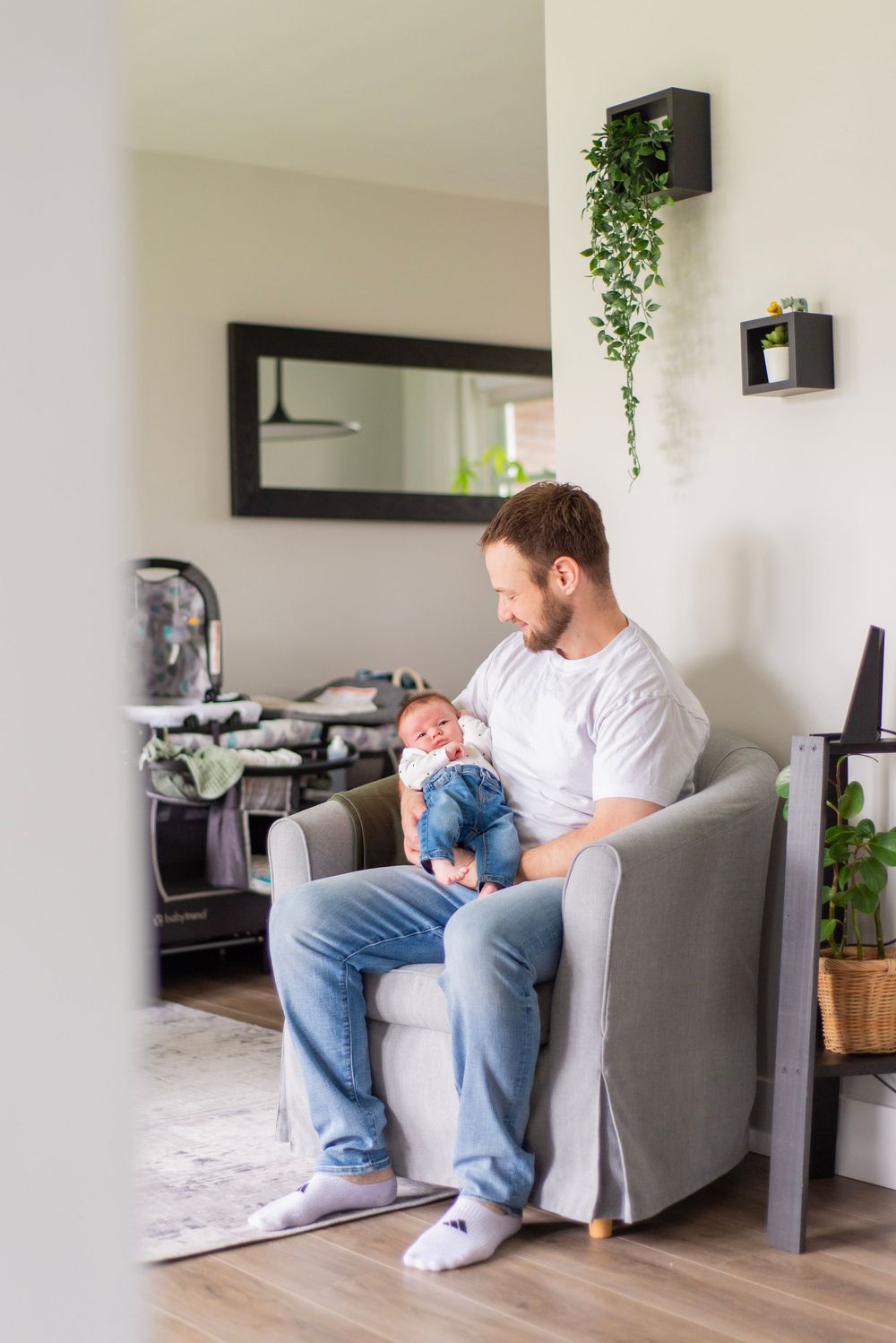 Parent sits in grey armchair holding infant in modern living room with hanging plants.