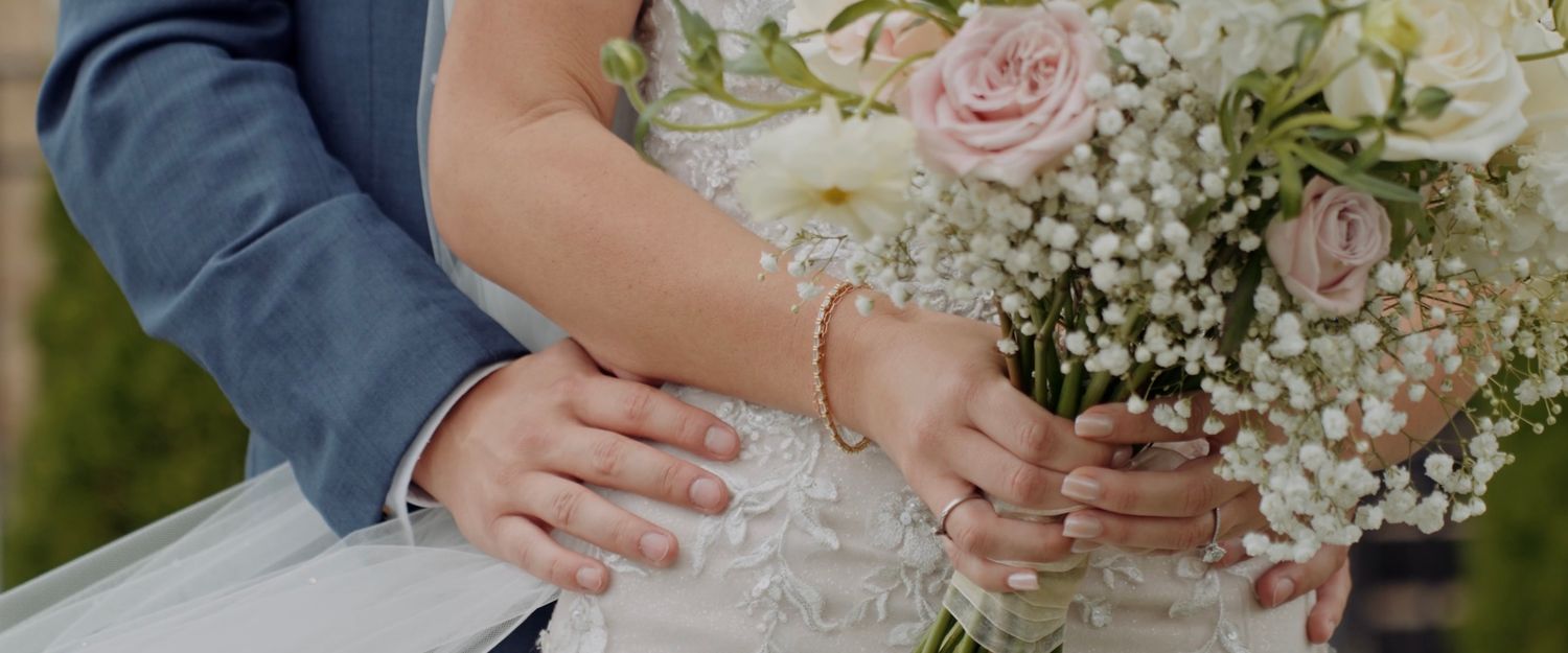 Close-up view of hands holding a delicate pink rose and baby's breath wedding bouquet against a formal suit.