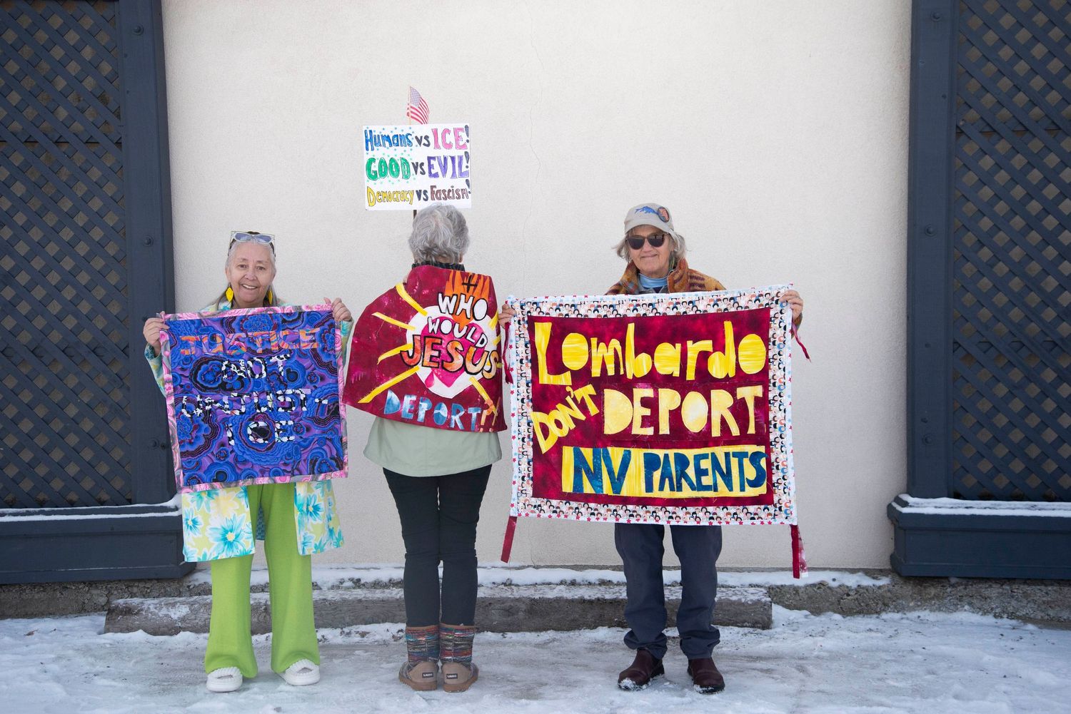 Three women holding colorful protest signs opposing deportation outside Governor's Mansion.
