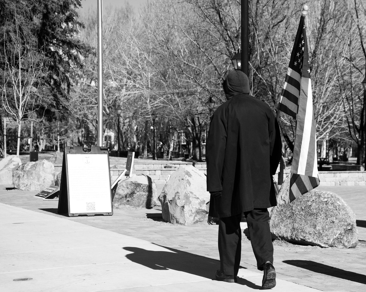 Hooded figure walks past American flag and boulders on sunny sidewalk in black and white photo.