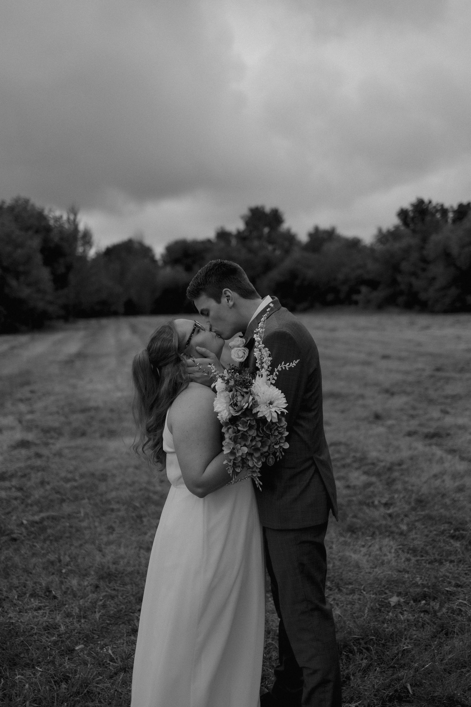 A romantic black and white wedding photo of a couple embracing in a field under dramatic cloudy skies.