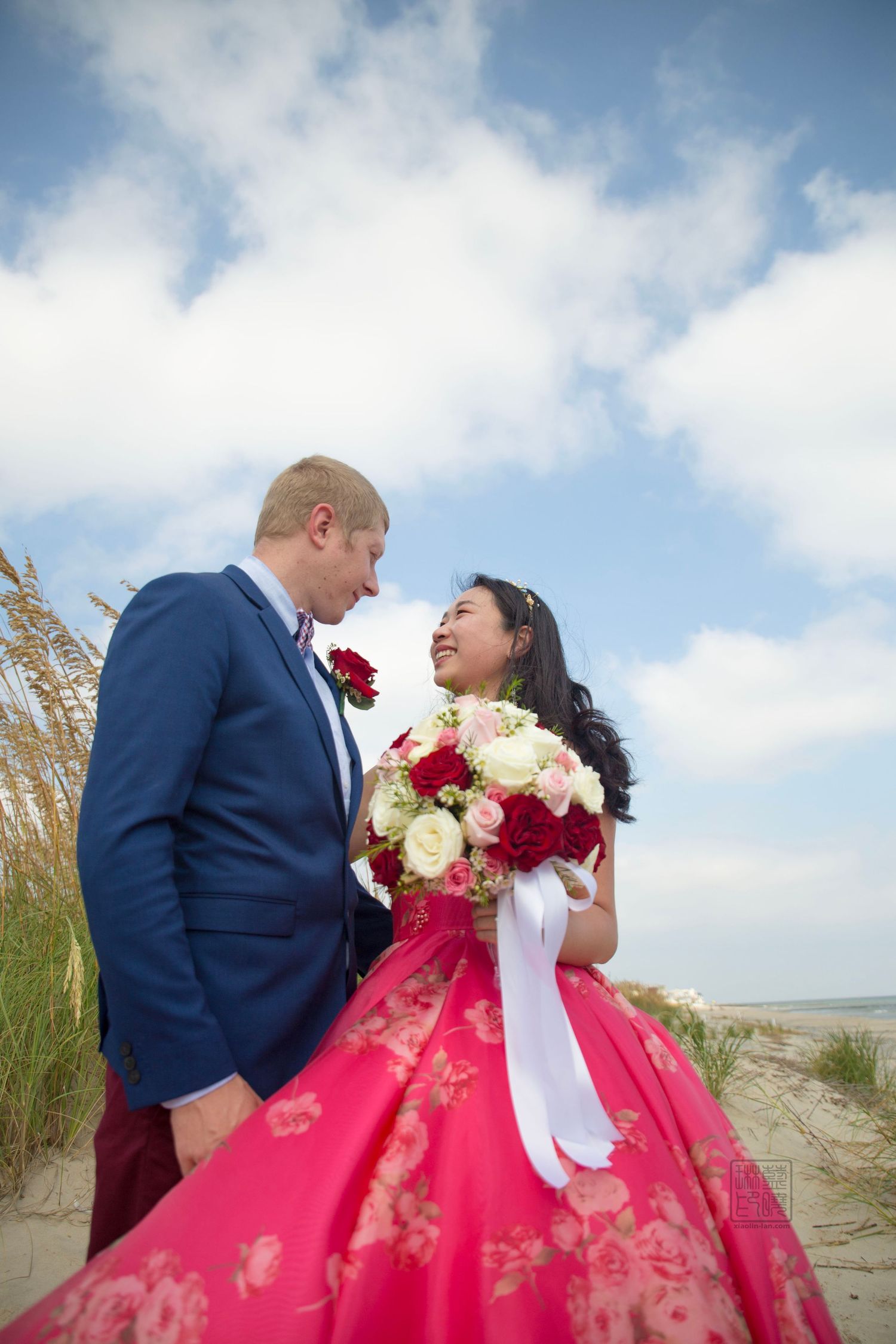 Couple poses for wedding photos on beach, bride wearing vibrant pink ballgown.